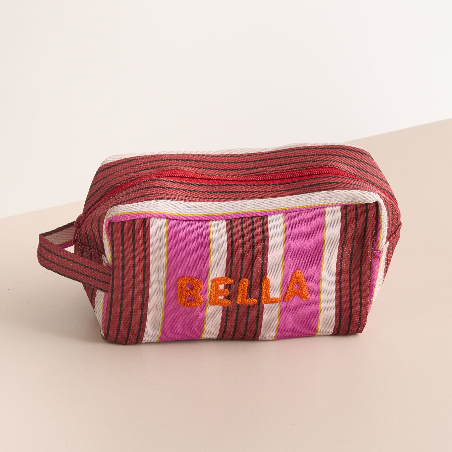 Eine gestreifte, bunte Kosmetik- oder Kulturtasche mit dem Aufdruck „Bella“ auf einer hellen Oberfläche.
