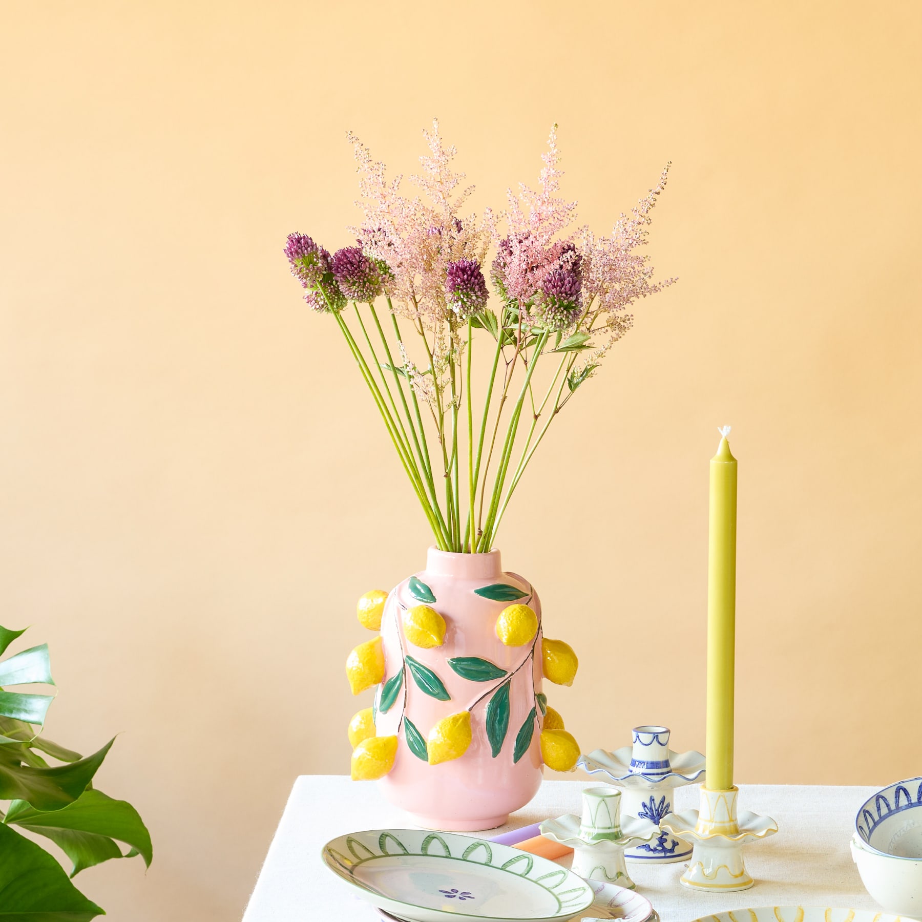 Eine rosa Keramikvase, verziert mit leuchtend gelben Zitronen und grünen Blättern, steht auf einer beigen Oberfläche vor einem schlichten Hintergrund.