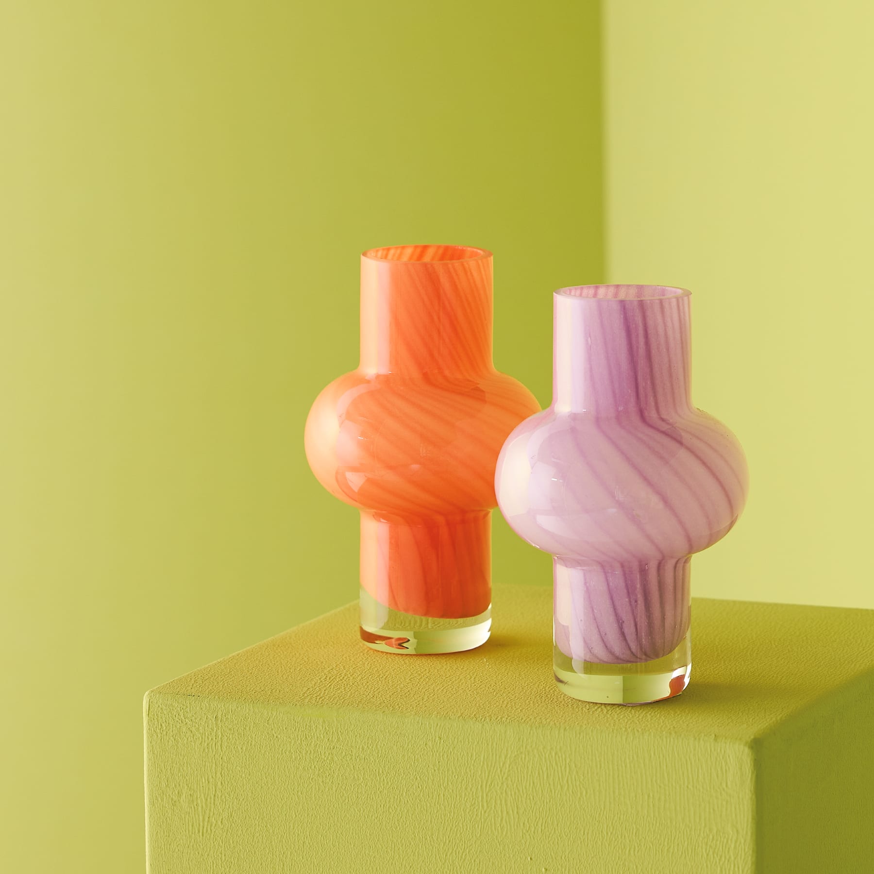 Eine leuchtend violette Alliumblüte steht hoch in einer orangefarbenen geometrischen Vase vor einem schlichten weißen Hintergrund.