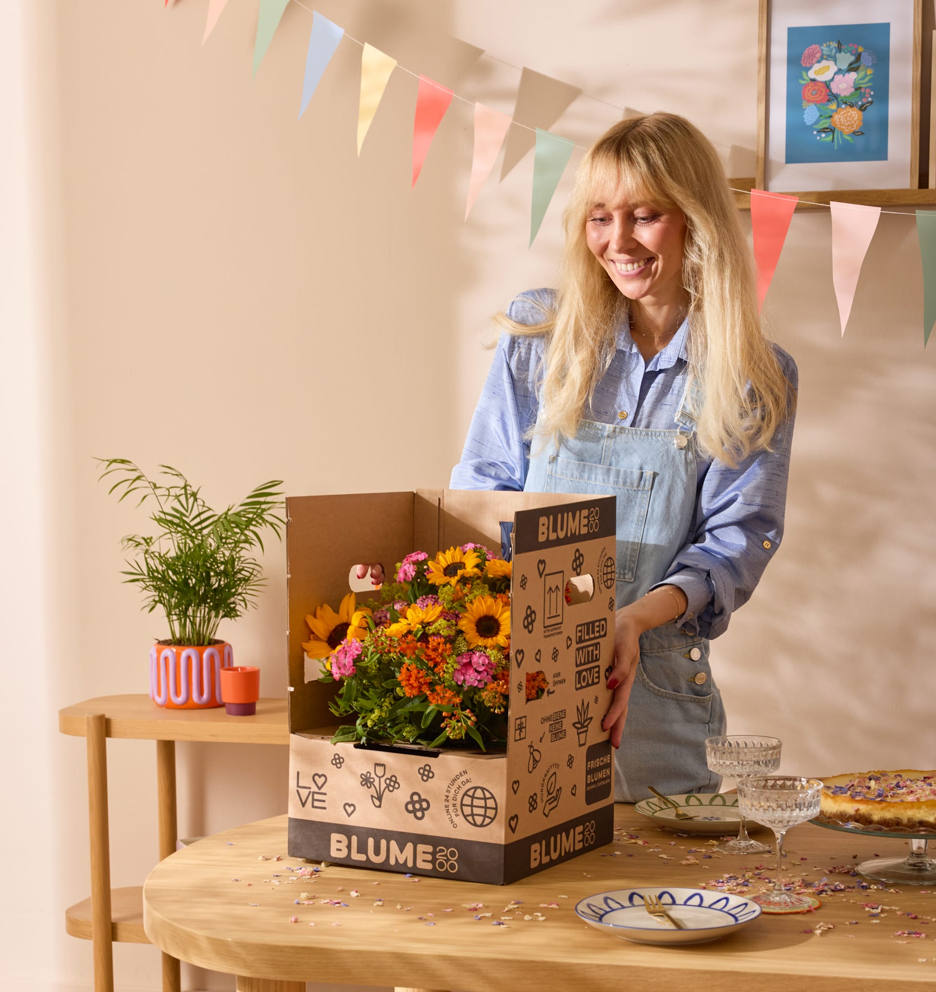 Eine lächelnde junge Frau in einer Jeansjacke steht hinter einem Holztisch, umgeben von einem bunten Blumenarrangement, einer Topfpflanze und einer Schachtel mit der Aufschrift „Blume“.