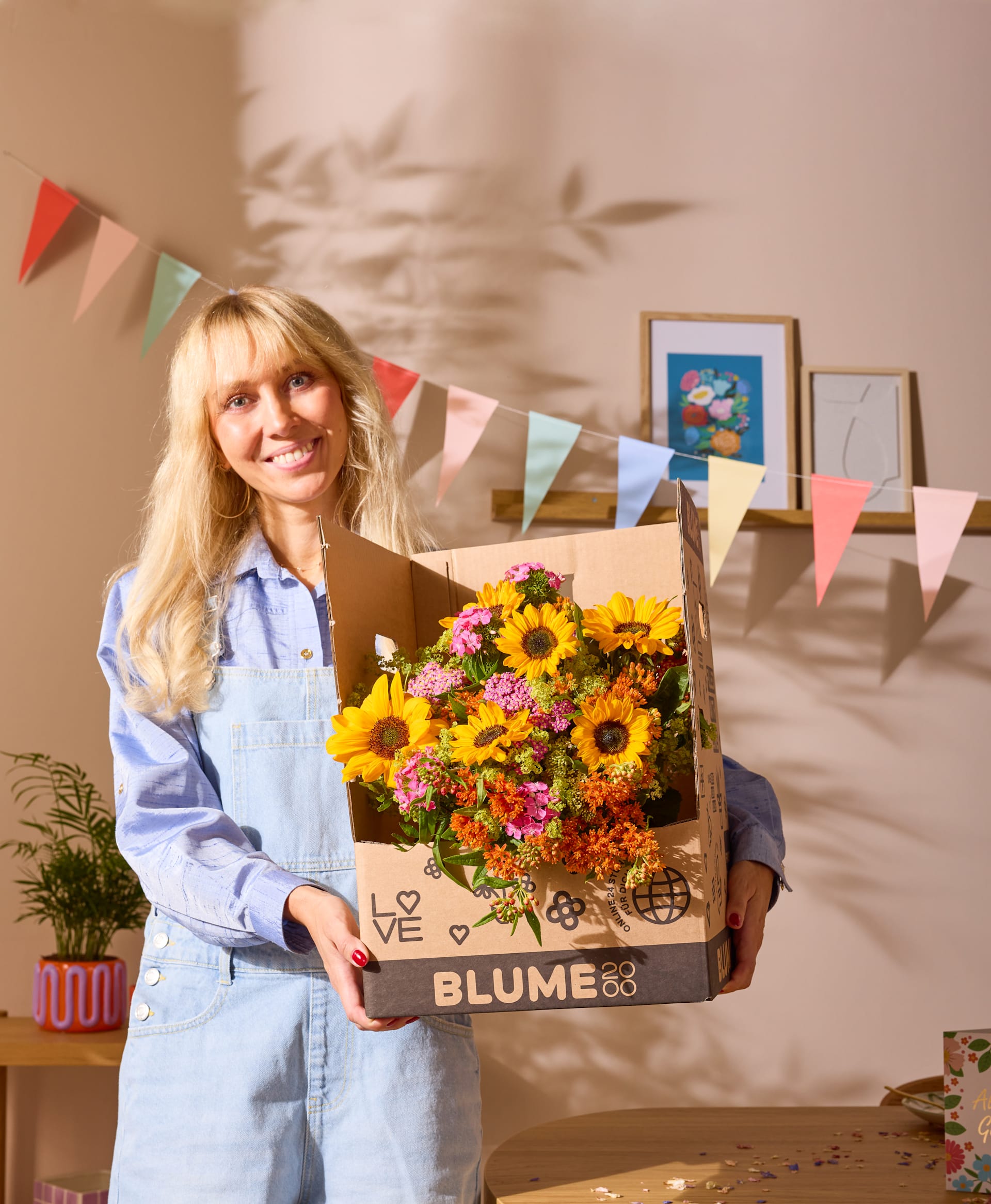 Eine lächelnde junge Frau mit einer Schachtel bunter Blumen steht in einem mit festlichen Fahnen und Kunstwerken geschmückten Raum.