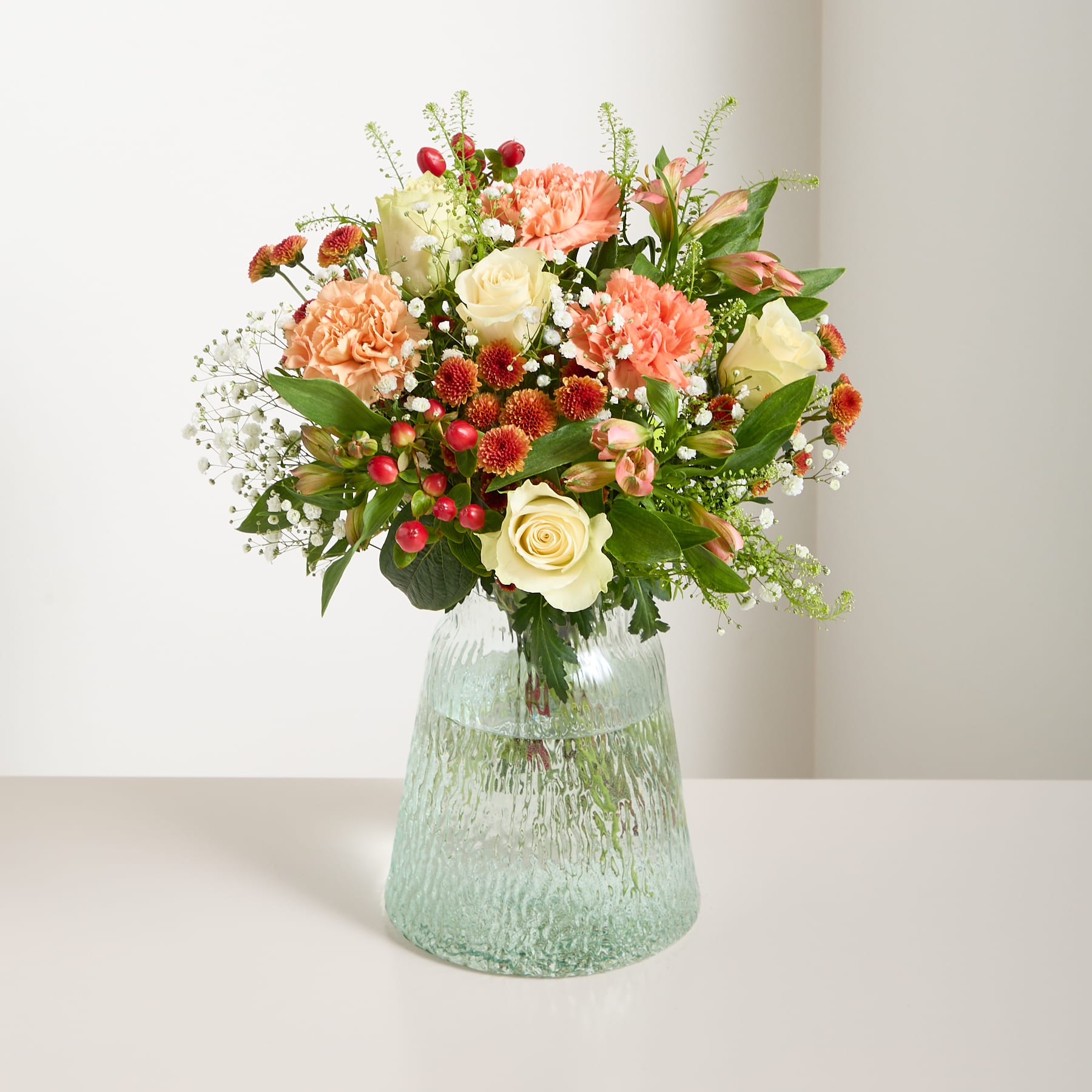 Ein leuchtender Strauß bunter Blumen, darunter gelbe Rosen, orangefarbene Nelken und verschiedene andere Blüten, wird vor einem schlichten weißen Hintergrund in der Hand einer Person gehalten.