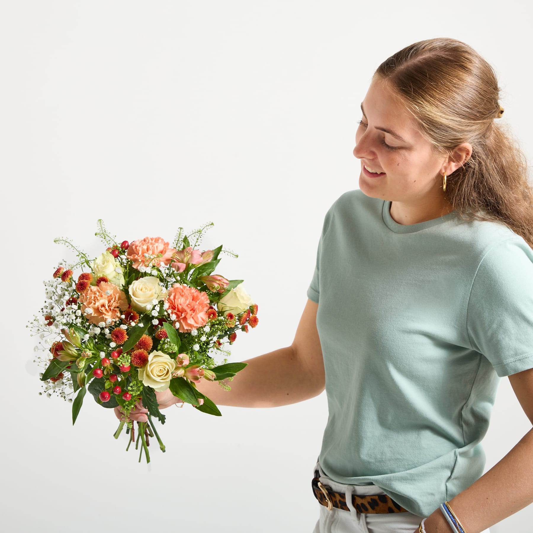 Ein leuchtender Strauß gelber Rosen, rosa Nelken und anderer bunter Blumen in einer grünen Glasvase steht auf einer hellen Fläche vor einem schlichten Hintergrund.