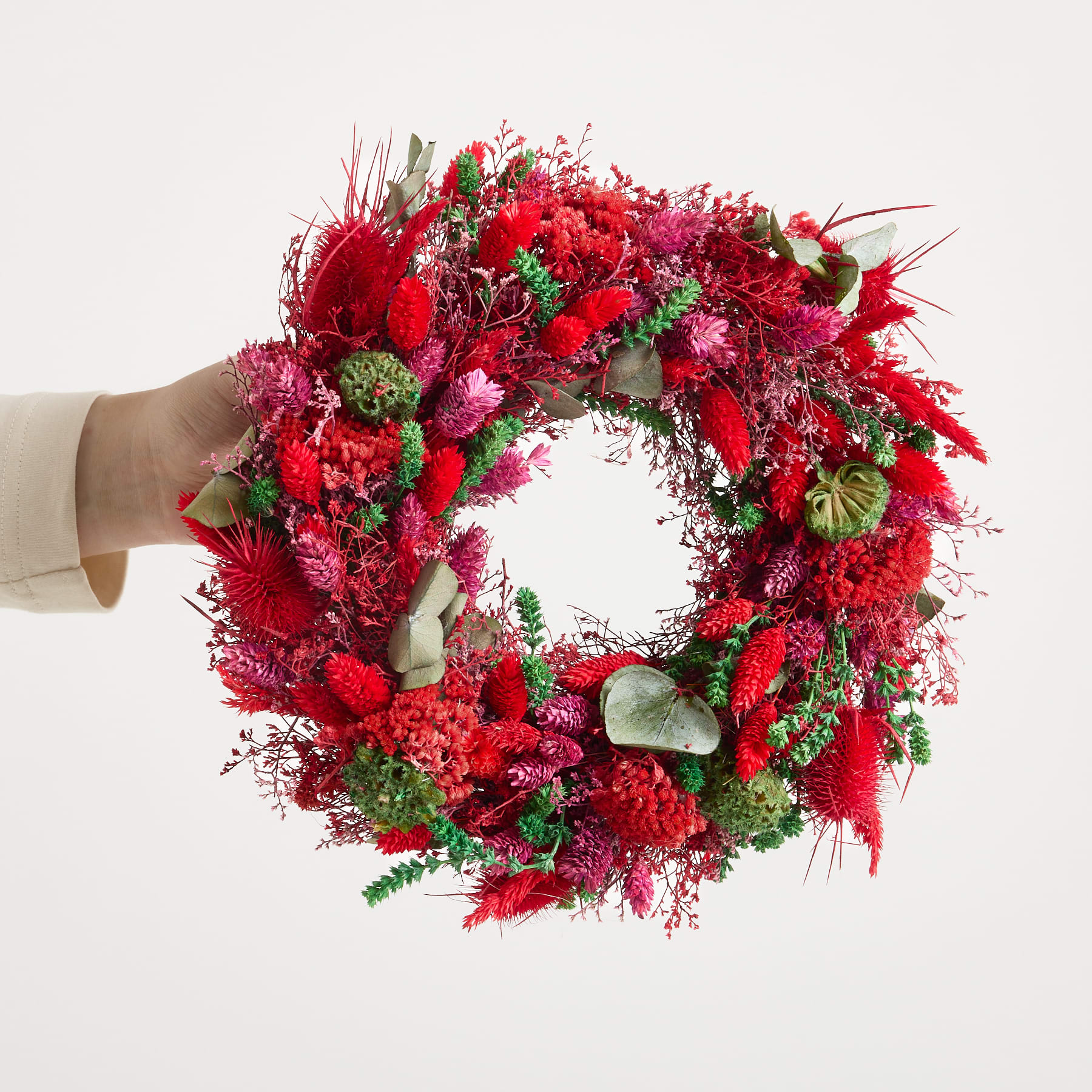 Ein leuchtend roter Blumenkranz wird in den Händen einer Person gehalten, die einen beigen Pullover vor einem schlichten, hellen Hintergrund trägt.