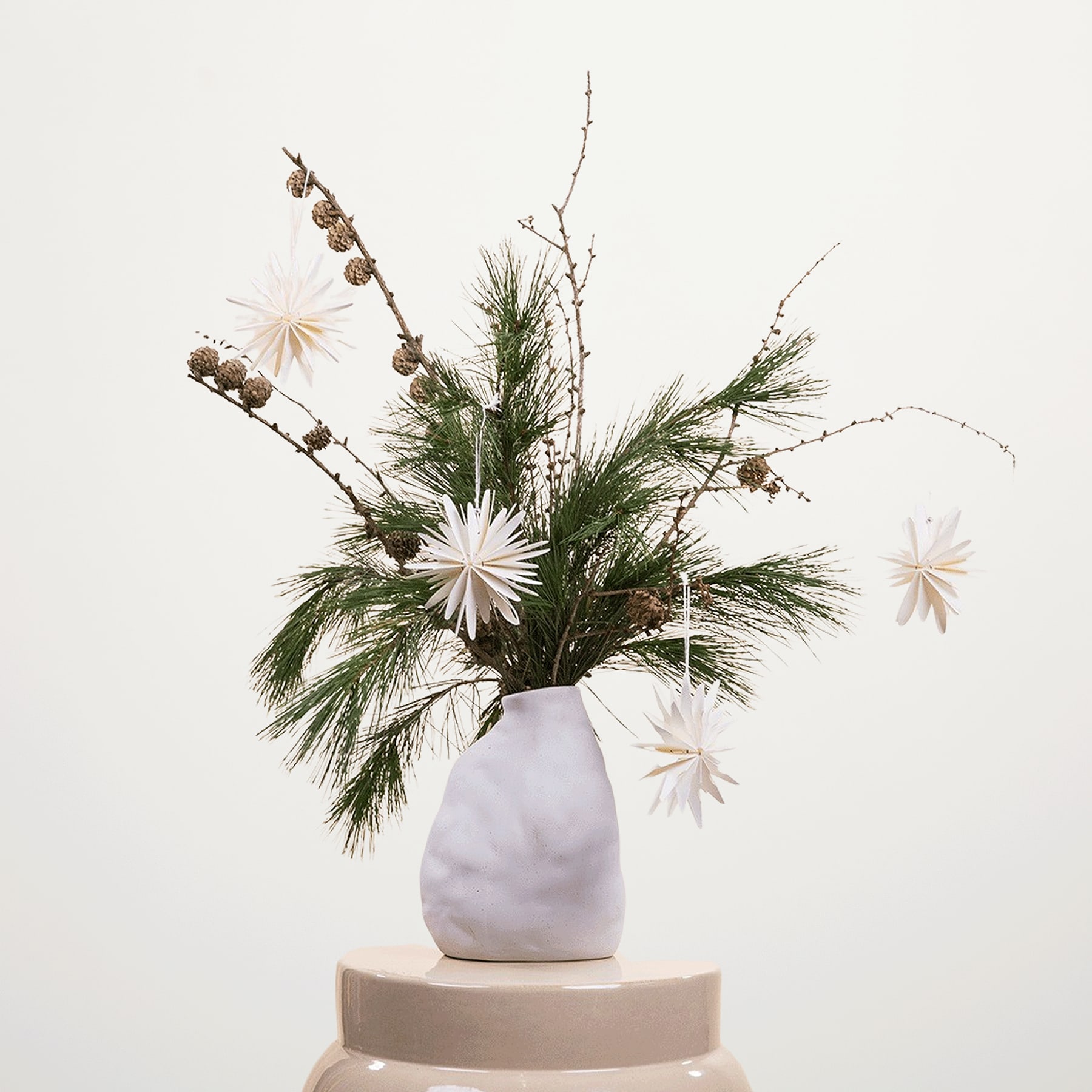 Eine weiße Keramikvase mit einem farbenfrohen Arrangement aus Tannenzweigen, getrockneten Blumen und weißen Gänseblümchen steht auf einem beigen Sockel vor einem schlichten, hellen Hintergrund.