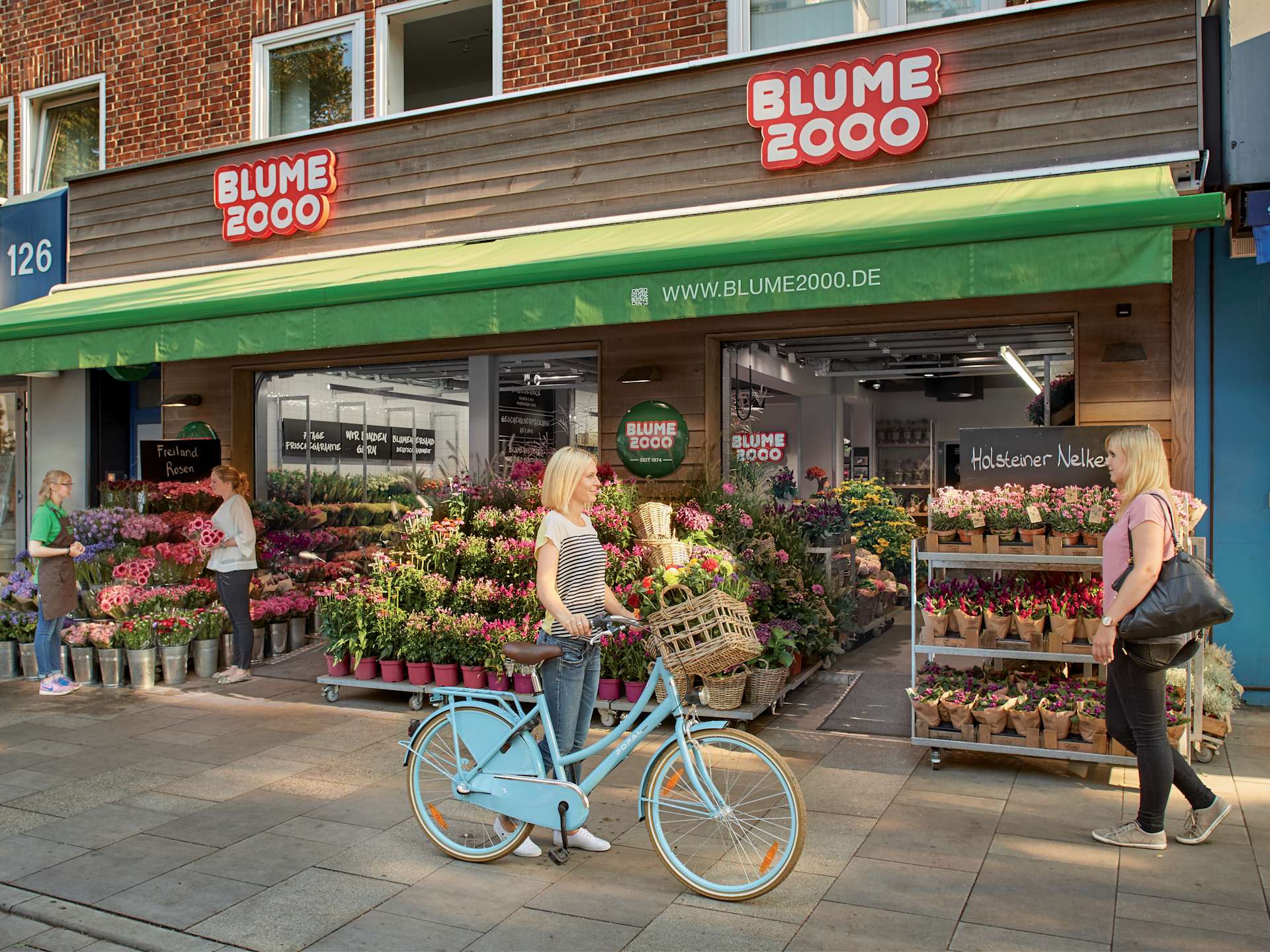 Ein lebhafter Blumenladen mit farbenfroher Fassade, umgeben von Menschen, die sich die Auswahl an Topfpflanzen und Blumen ansehen, und einem blauen Fahrrad, das im Vordergrund parkt.