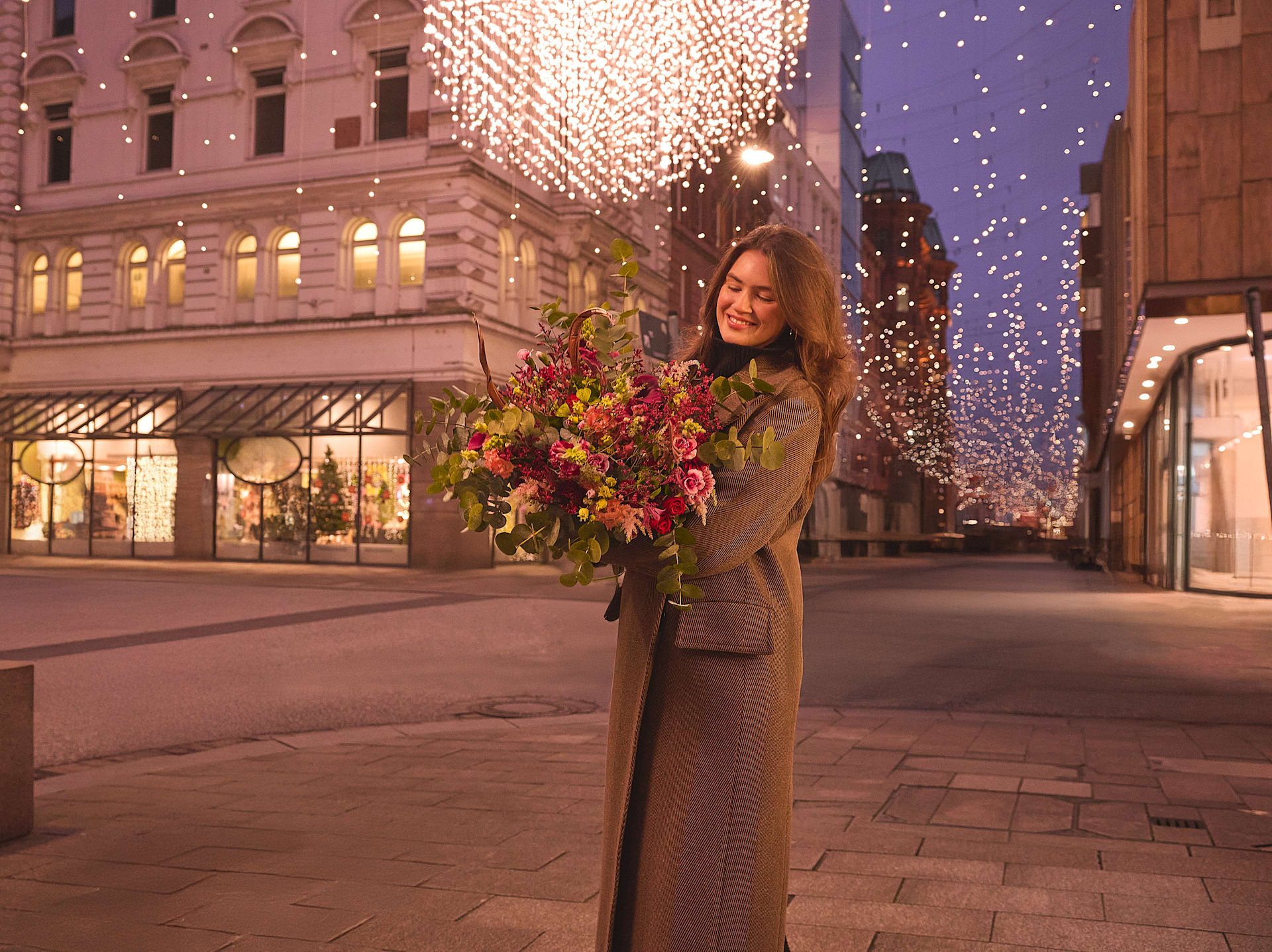 Eine Frau in einem beigen Mantel steht nachts auf einer Stadtstraße und hält einen Strauß bunter Blumen in der Hand. Im Hintergrund schmücken festliche Lichter und Dekorationen die Gebäude.
