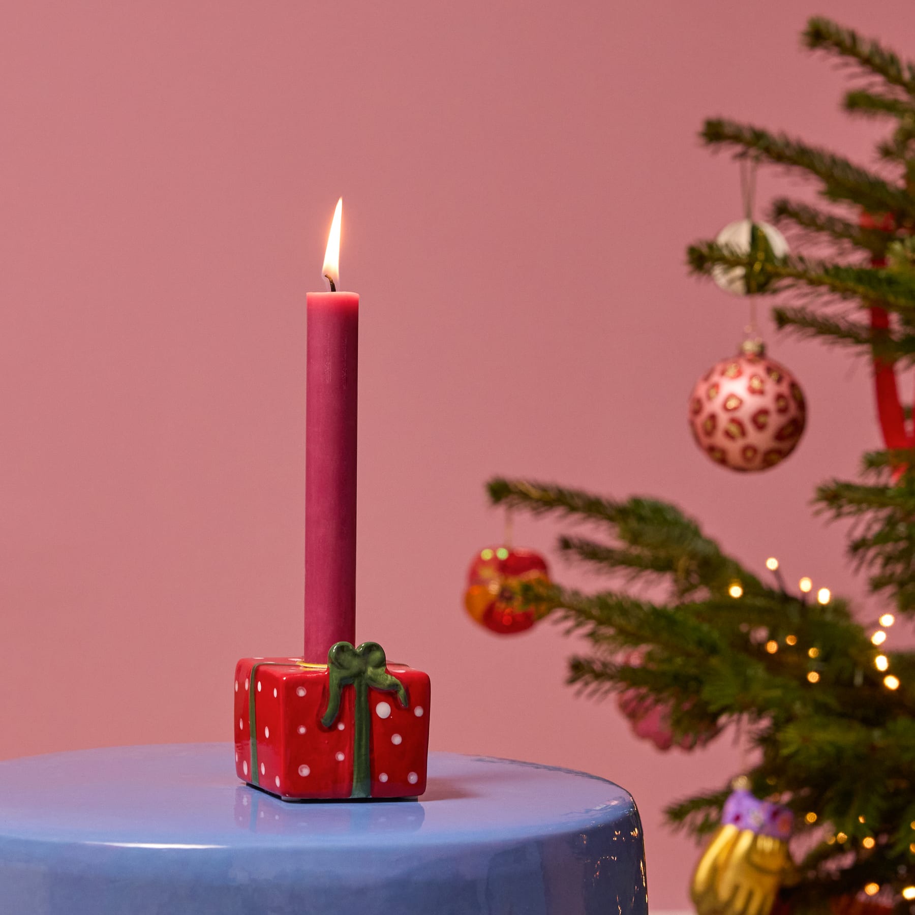 Eine rote Geschenkbox mit weißen Punkten und einer grünen Schleife steht auf einer Oberfläche und trägt eine große weiße Kerze.