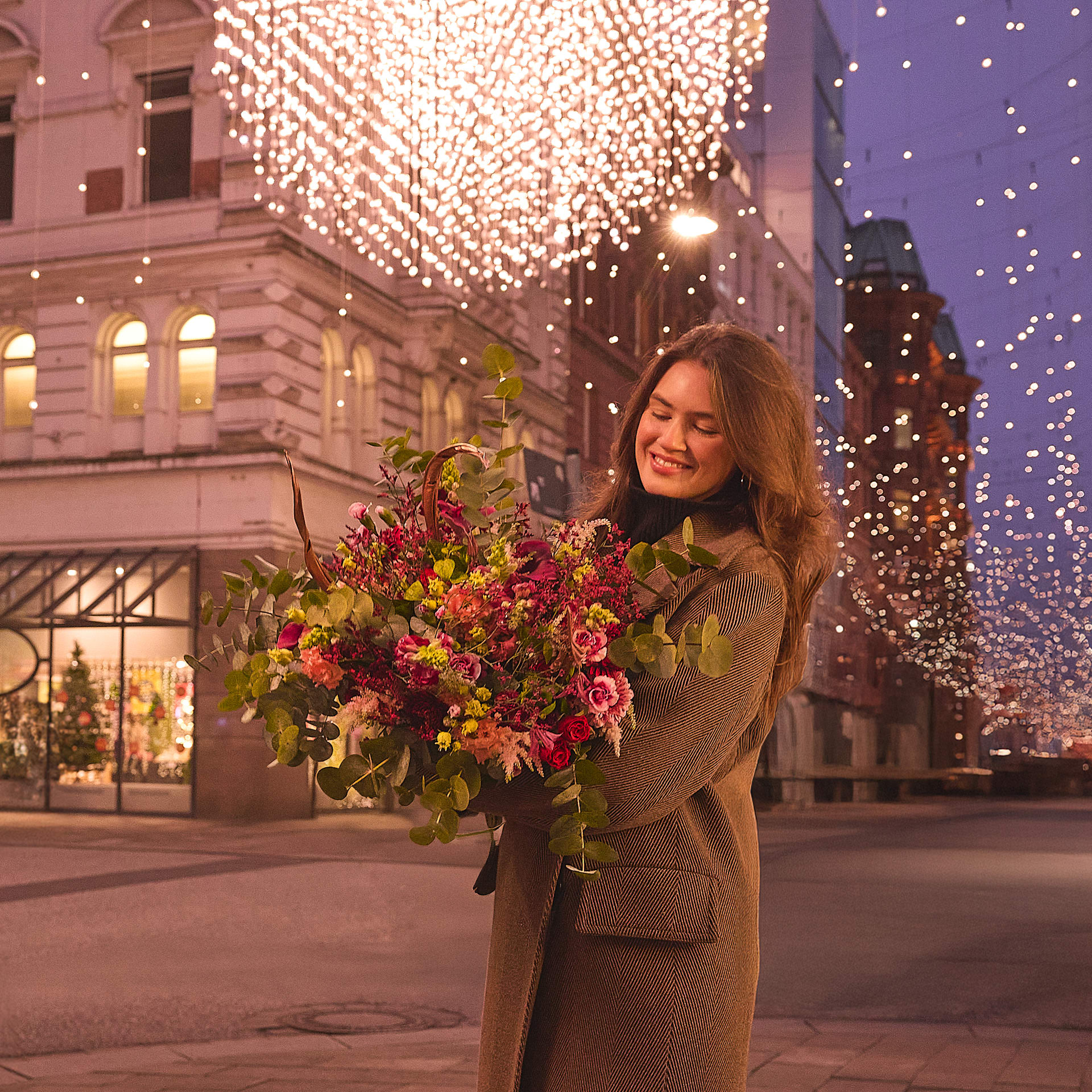Eine Frau in einem beigen Mantel steht vor einem Gebäude mit einem großen, beleuchteten Kronleuchter und hält einen Strauß bunter Blumen, während um sie herum Schneeflocken fallen.