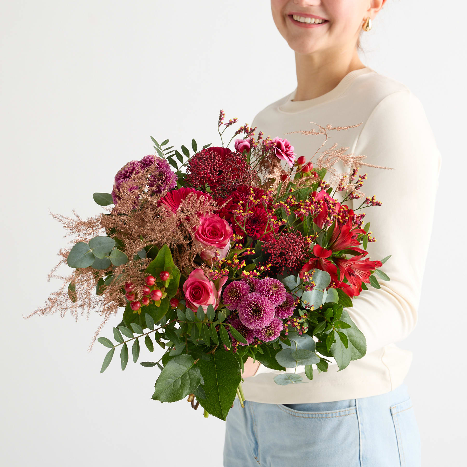 Eine lächelnde junge Frau hält einen farbenfrohen Strauß roter, rosa und violetter Blumen vor einem schlichten weißen Hintergrund.