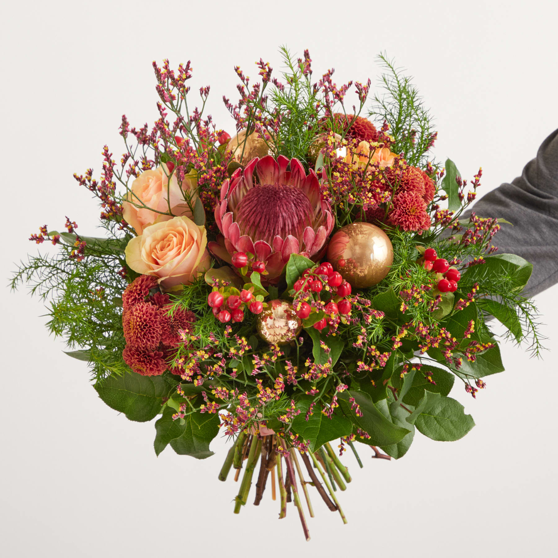Ein farbenfroher Strauß aus verschiedenen herbstlich gefärbten Blumen, darunter Proteen, Rosen und Beeren, in den Händen einer Person vor einem schlichten weißen Hintergrund.