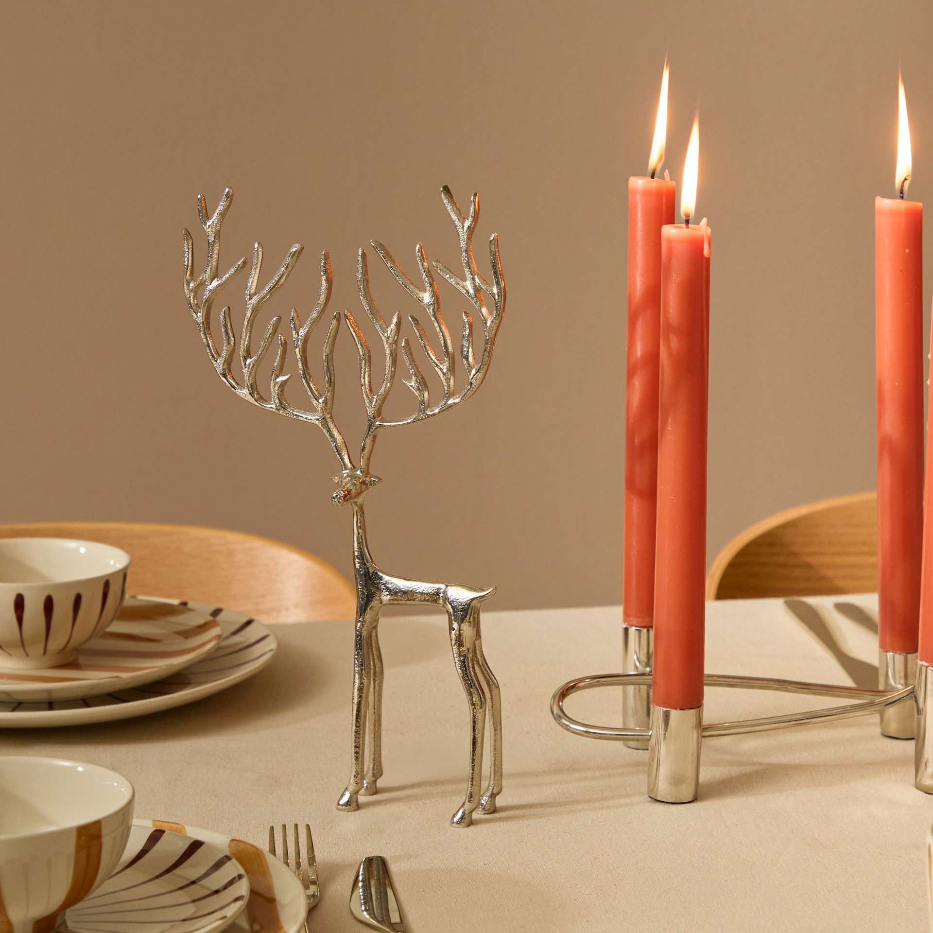 Auf einem Tisch steht ein silberner, hirschförmiger Kerzenhalter, umgeben von Tellern und brennenden roten Kerzen vor einem warmtonigen Hintergrund.