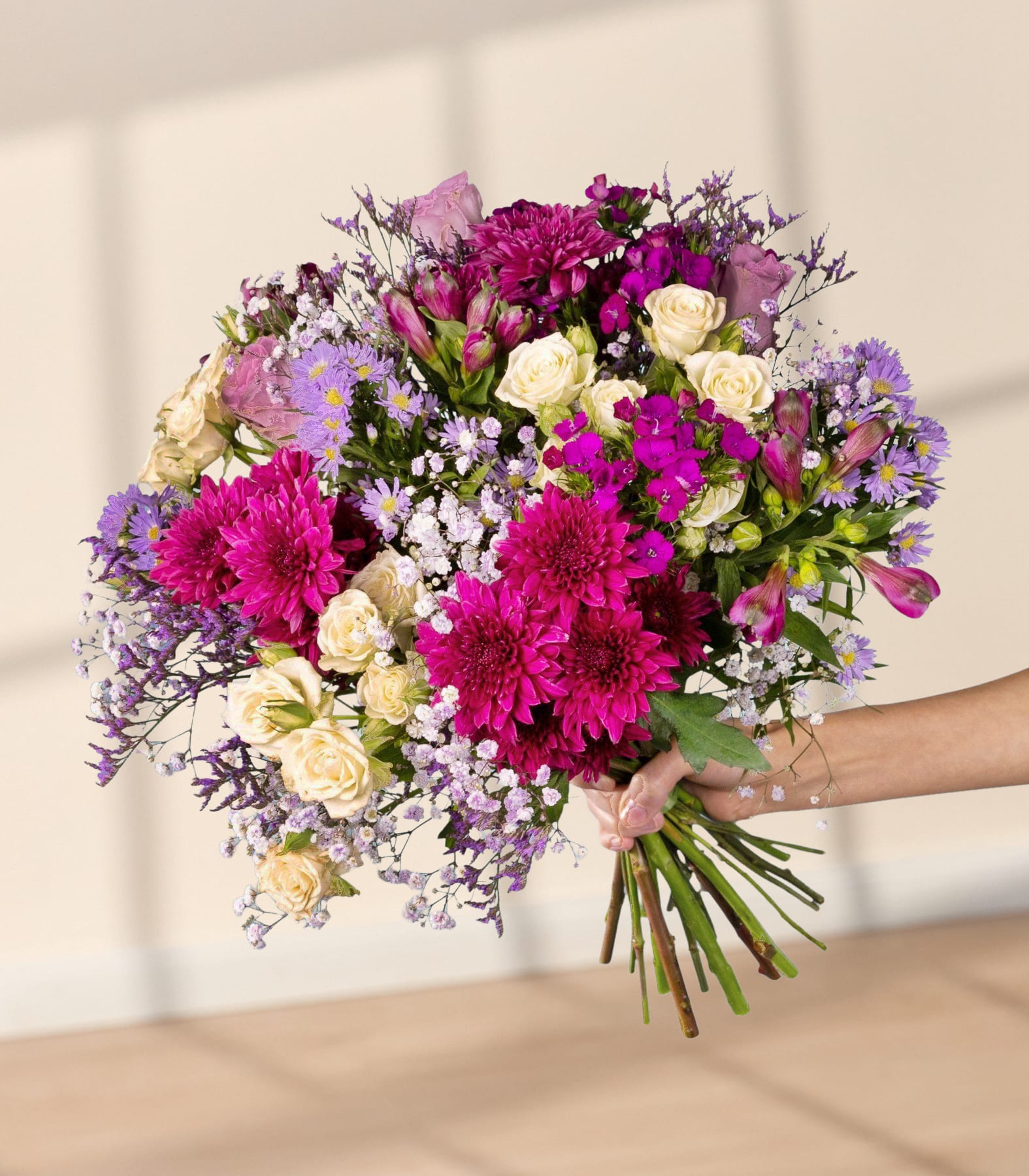 Ein lebendiger Strauß verschiedener bunter Blumen, darunter rosa und violette Chrysanthemen, weiße Rosen und zarte violette und weiße Blumen, die eine Person vor einem schlichten weißen Hintergrund in den Händen hält.