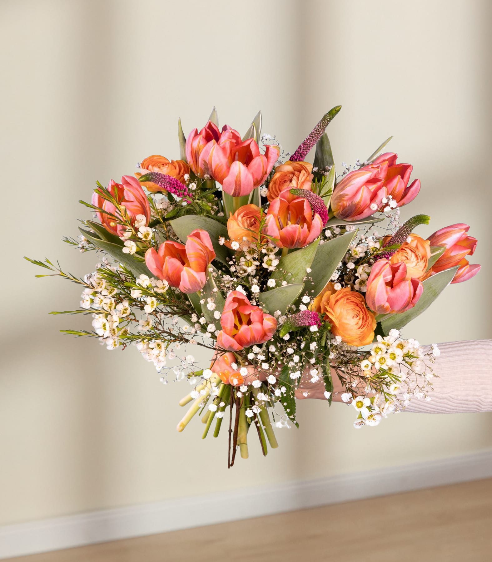 Ein leuchtender Strauß rosa und orangefarbener Tulpen, umgeben von Grün und Schleierkraut, wird vor einem schlichten weißen Hintergrund in den Händen einer Person gehalten.