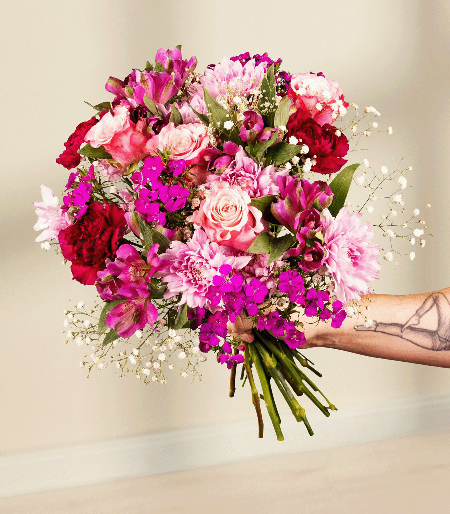 Ein leuchtender Strauß rosa, roter und violetter Blumen, darunter Rosen, Nelken und Schleierkraut, wird von einer tätowierten Hand einer Person vor einem schlichten blauen Hintergrund gehalten.