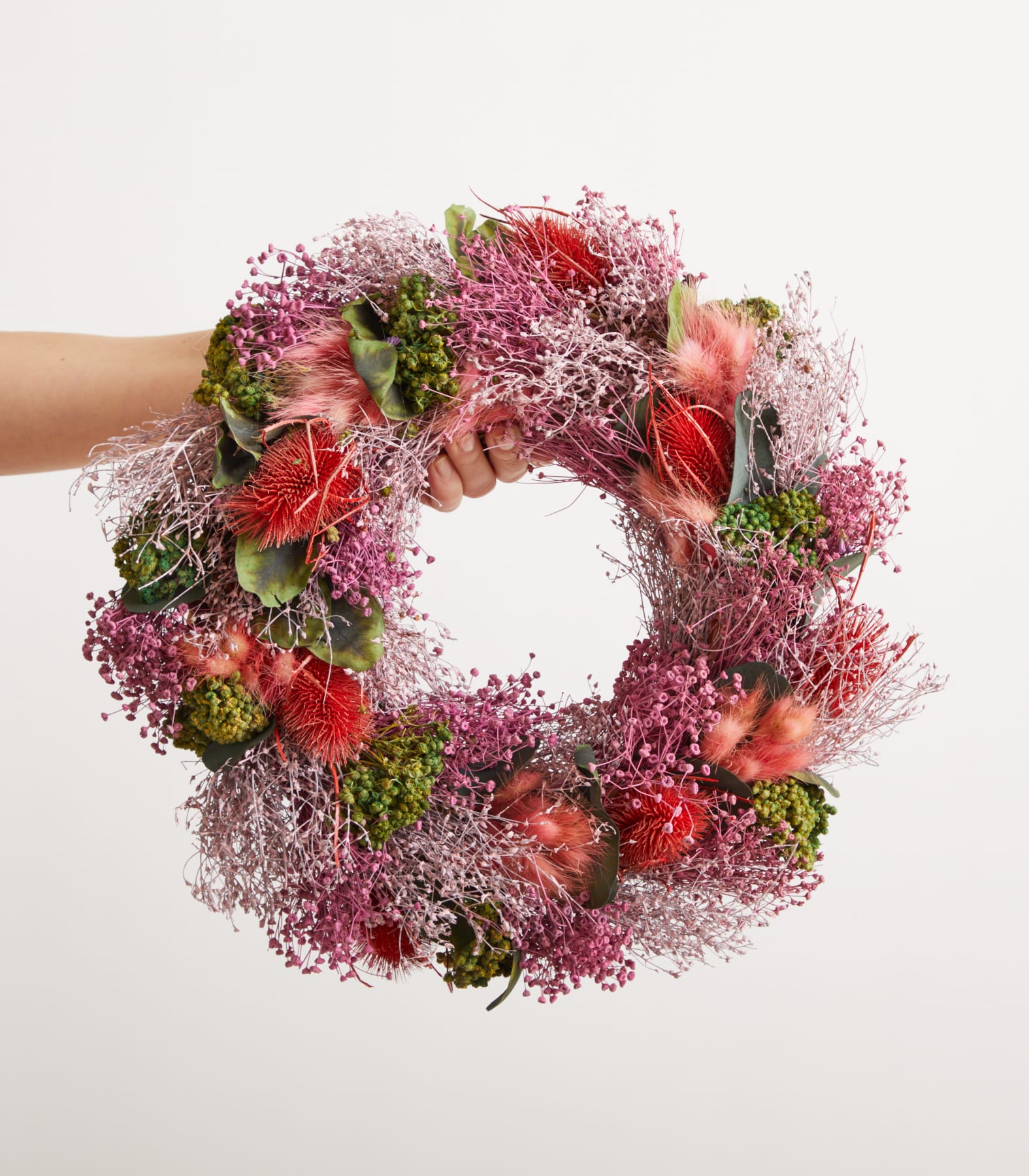 Eine Person hält vor einem schlichten weißen Hintergrund einen lebendigen und farbenfrohen Blumenkranz aus verschiedenen Blumen und Grünpflanzen in den Händen.