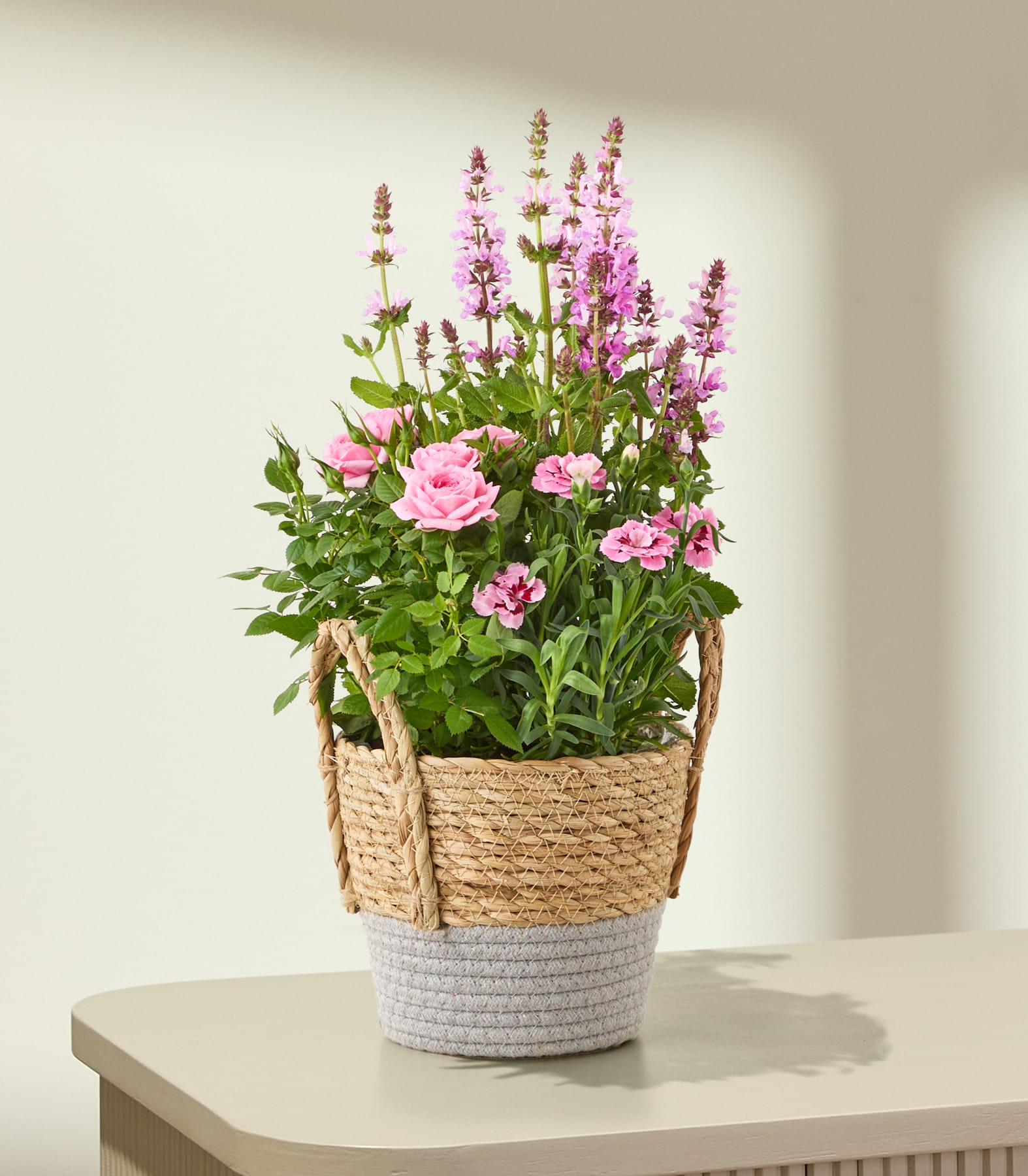 Ein farbenfrohes Arrangement aus rosa Blumen und violetten Blüten in einem geflochtenen Korb vor einem schlichten weißen Hintergrund.