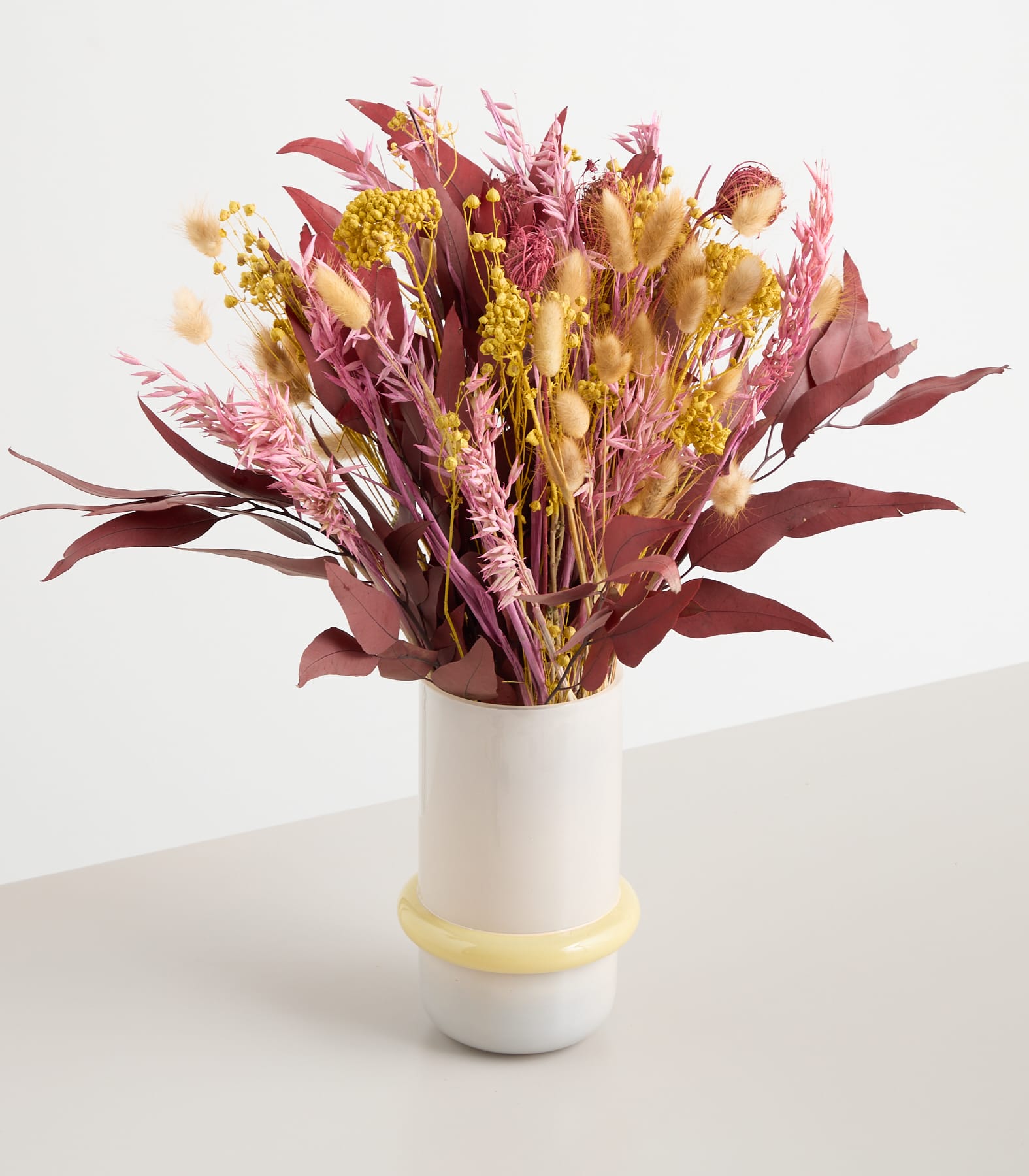Ein farbenfroher Strauß getrockneter Blumen in Rosa-, Gelb- und Burgundertönen wird in einer einfachen weißen Vase auf einem schlichten Hintergrund präsentiert.