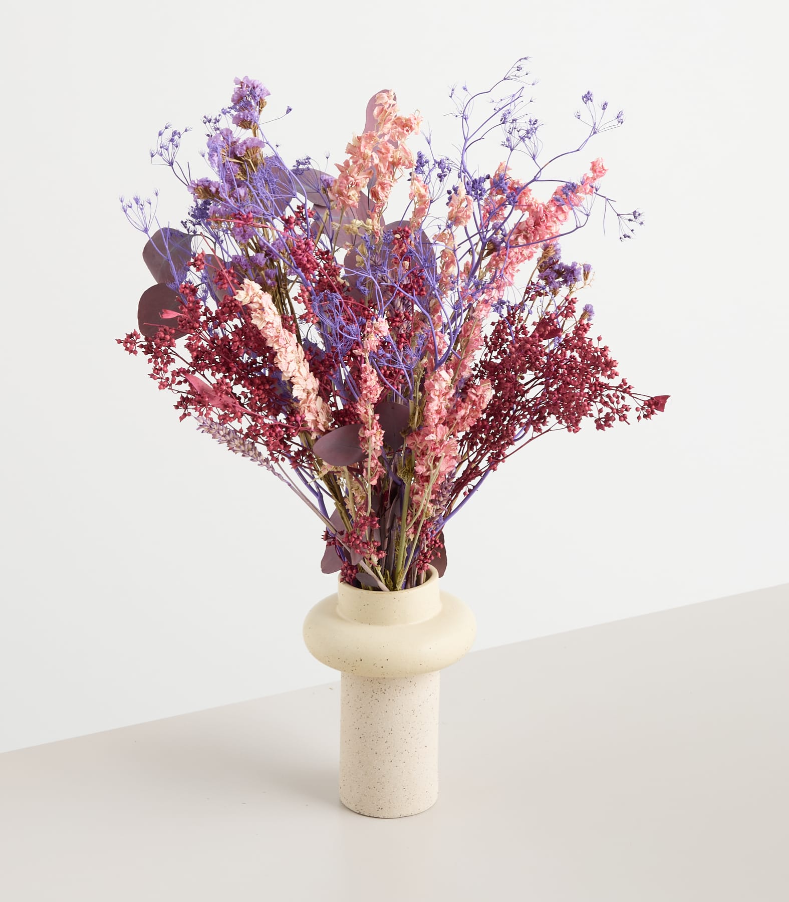 Ein farbenfroher Strauß getrockneter Blumen in verschiedenen Lila-, Rosa- und Rottönen steht auf einem schlichten weißen Hintergrund und schafft eine auffällige und optisch ansprechende Komposition.