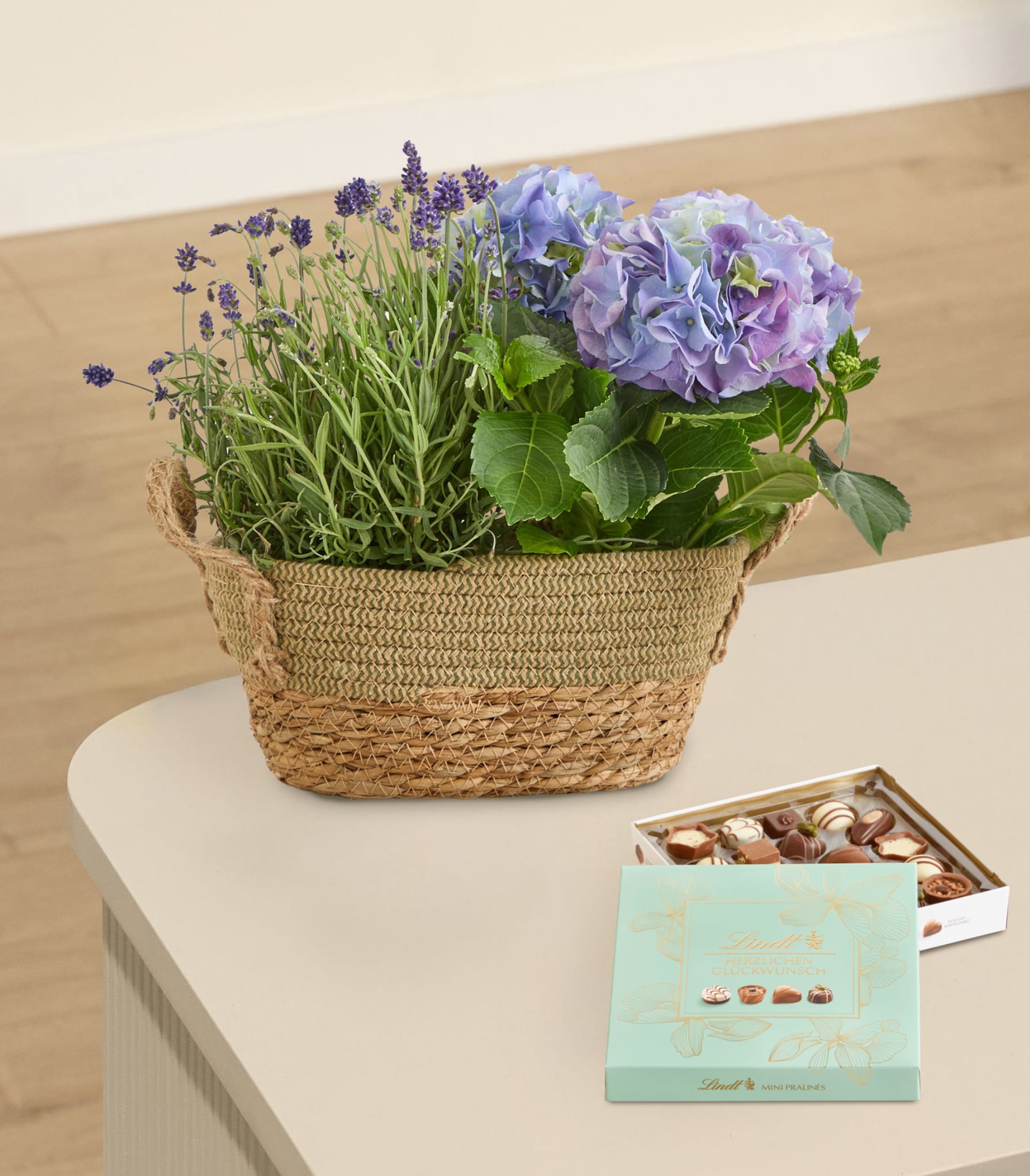 Auf einer hellen Oberfläche steht ein geflochtener Korb mit leuchtend violetten und grünen Blütenpflanzen, daneben eine kleine Schachtel Pralinen.