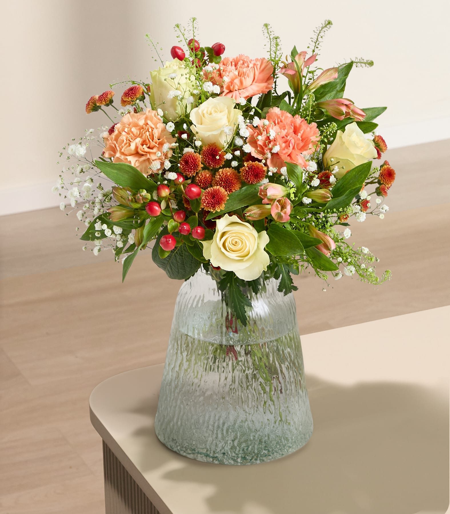 Ein leuchtender Strauß bunter Blumen, darunter gelbe Rosen, orangefarbene Nelken und verschiedene andere Blüten, wird vor einem schlichten weißen Hintergrund in der Hand einer Person gehalten.