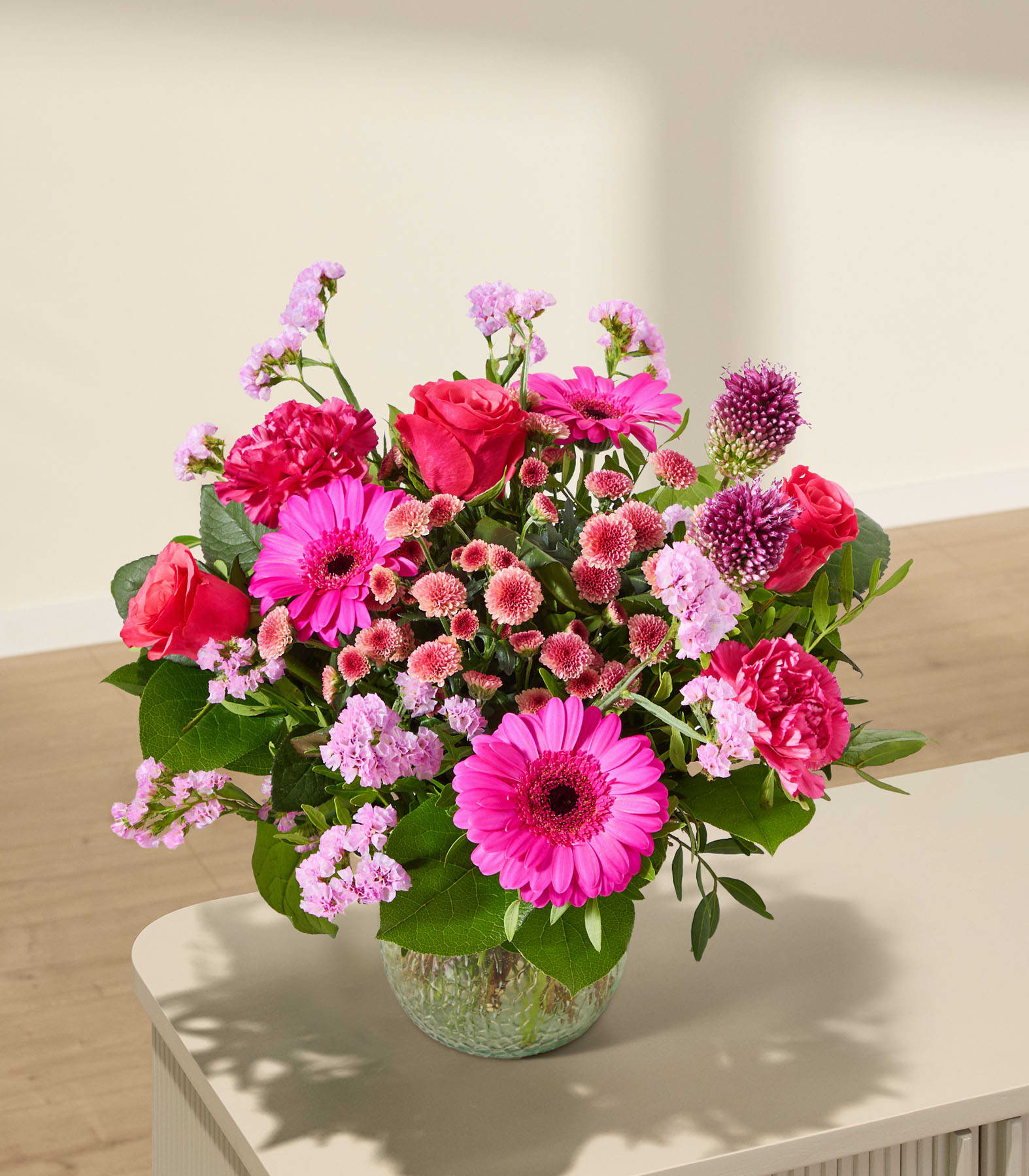 Ein leuchtender Strauß rosa und roter Blumen steht in einer strukturierten grünen Vase auf einer hellen Oberfläche, im Hintergrund eine sanft beleuchtete, neutrale Wand.