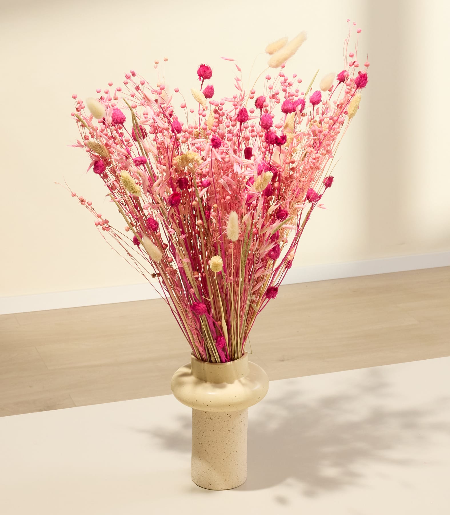 Ein Strauß aus leuchtend pinken und roten Trockenblumen, arrangiert in einer schlichten Keramikvase vor einem einfarbigen, hellen Hintergrund.