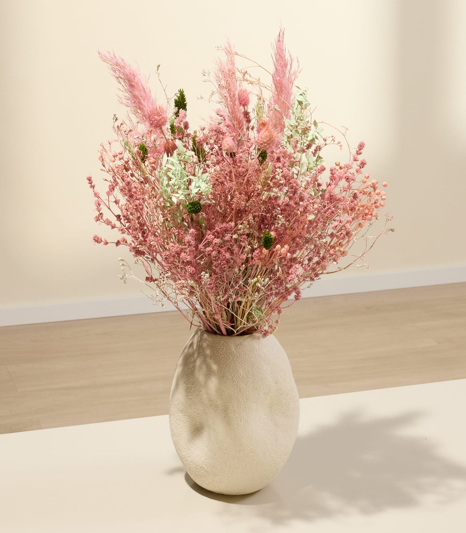 Eine Vase, gefüllt mit einem farbenfrohen Strauß aus rosa und weißen Trockenblumen, steht auf einer Holzoberfläche vor einem schlichten Hintergrund.