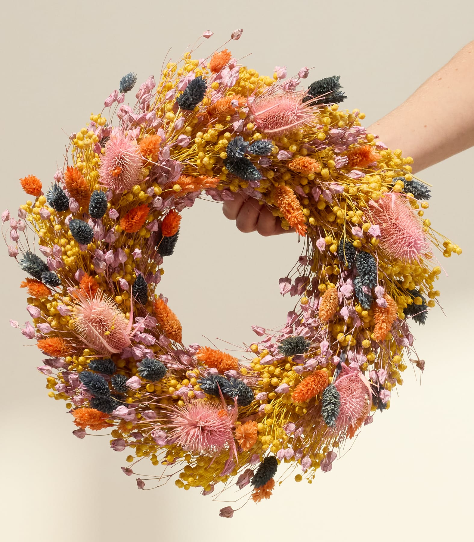 Ein lebendiger, farbenfroher Kranz aus verschiedenen Trockenblumen und -blättern, der von einer Person in den Händen gehalten wird, vor einem neutralen Hintergrund.