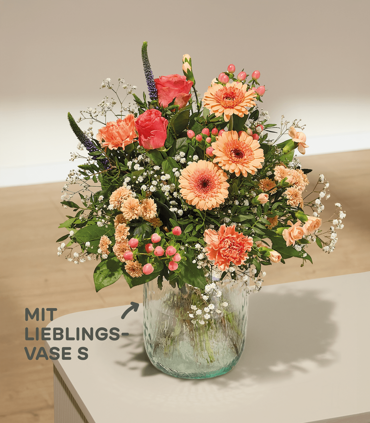 Ein lebhafter Strauß bunter Blumen, darunter orangefarbene Gerbera, rosa Nelken und weißes Schleierkraut, arrangiert in einer Glasvase vor einem weichen, verschwommenen Hintergrund.