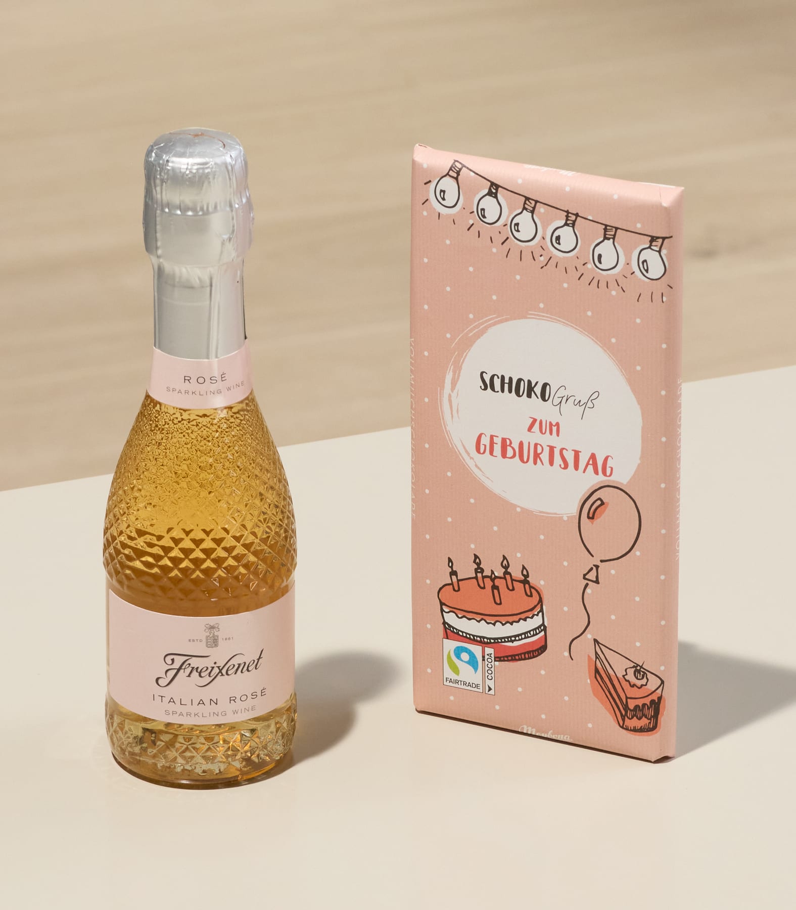 Auf einer Holzoberfläche stehen eine Flasche Sekt und eine rosa Geschenkbox mit einem Geburtstagskuchenmotiv.
