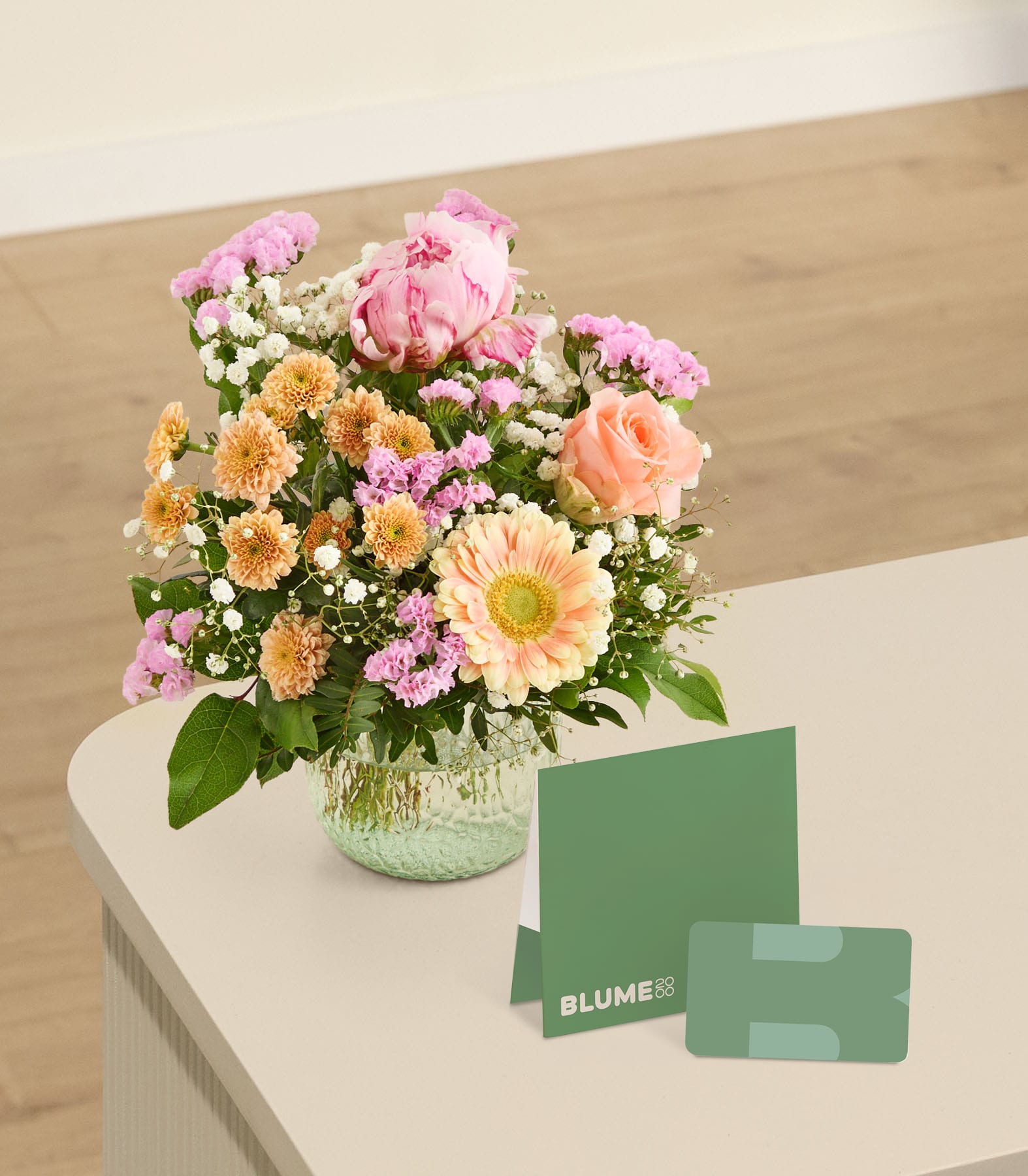 Auf einer Holzoberfläche steht ein üppiger Strauß aus rosa Pfingstrosen, gelben Gänseblümchen und anderen farbenfrohen Blumen, daneben eine grüne Karte mit dem Aufdruck „Blume“.