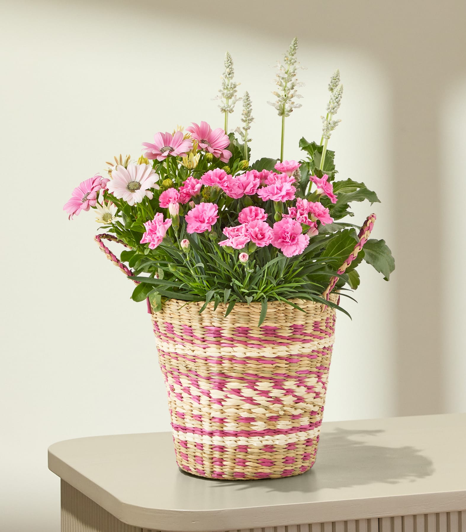 Auf einer hellen Fläche steht ein geflochtener Korb, gefüllt mit rosa und weißen Blumen; im Hintergrund ist eine sanft beleuchtete, strukturierte Wand zu sehen.