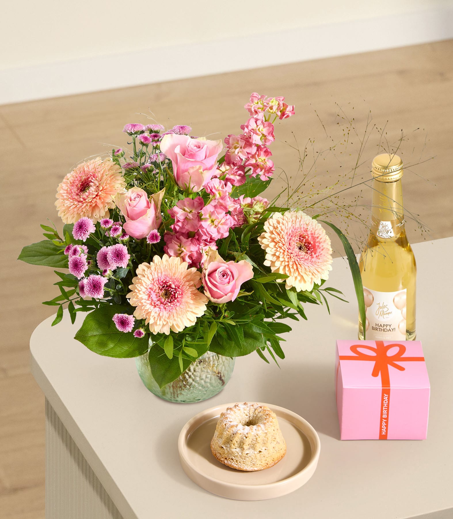 Auf einem Tisch vor einem leuchtenden Strauß rosa und pfirsichfarbener Blumen stehen eine rosa Geschenkbox, eine Flasche Sekt und ein kleiner Kuchen; im Hintergrund ist eine helle Wand zu sehen.