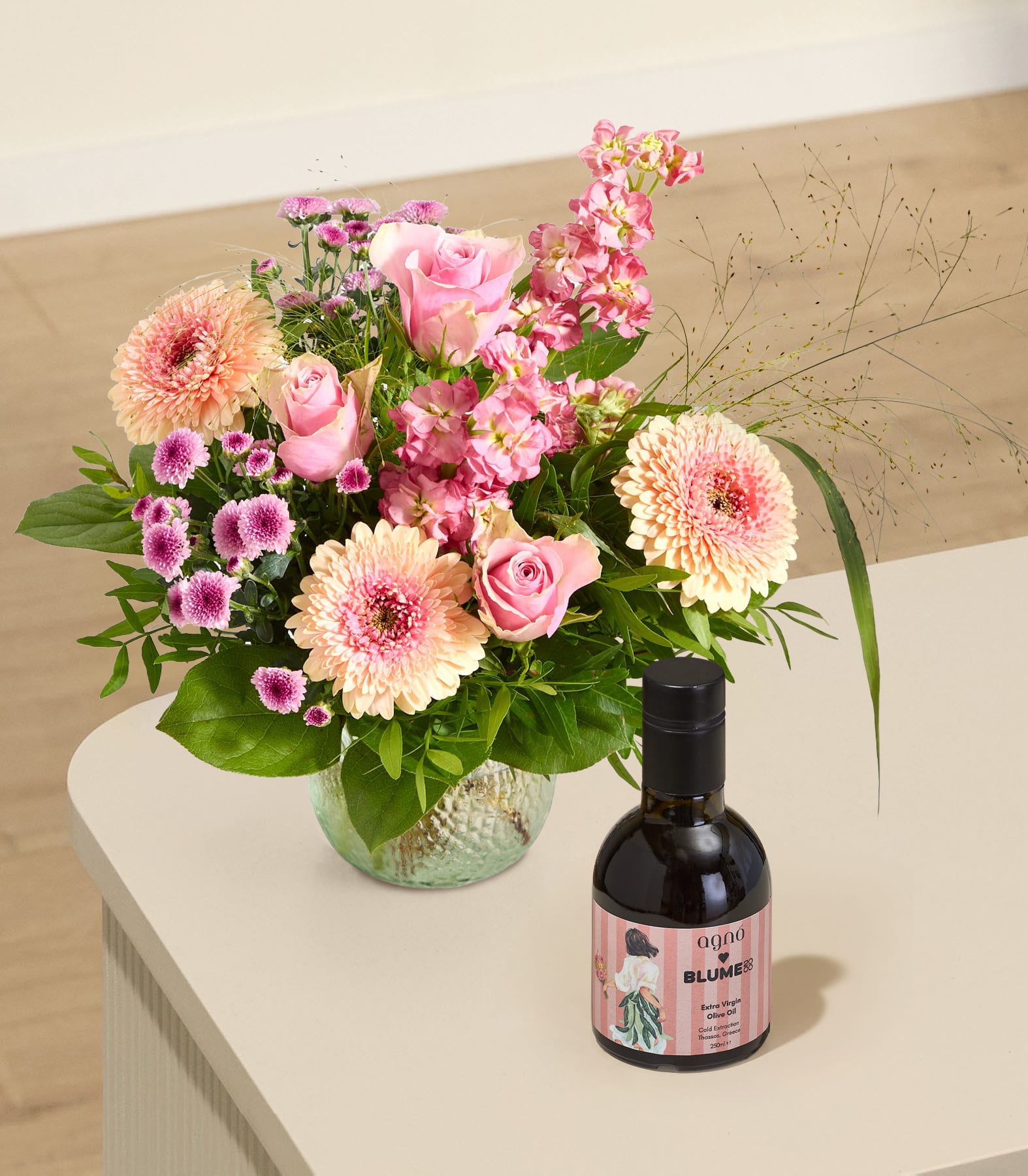 Auf einem hellen Tisch steht ein Strauß rosa und pfirsichfarbener Blumen in einer Glasvase neben einer Flasche Olivenöl; im Hintergrund ist ein Zimmer verschwommen zu sehen.