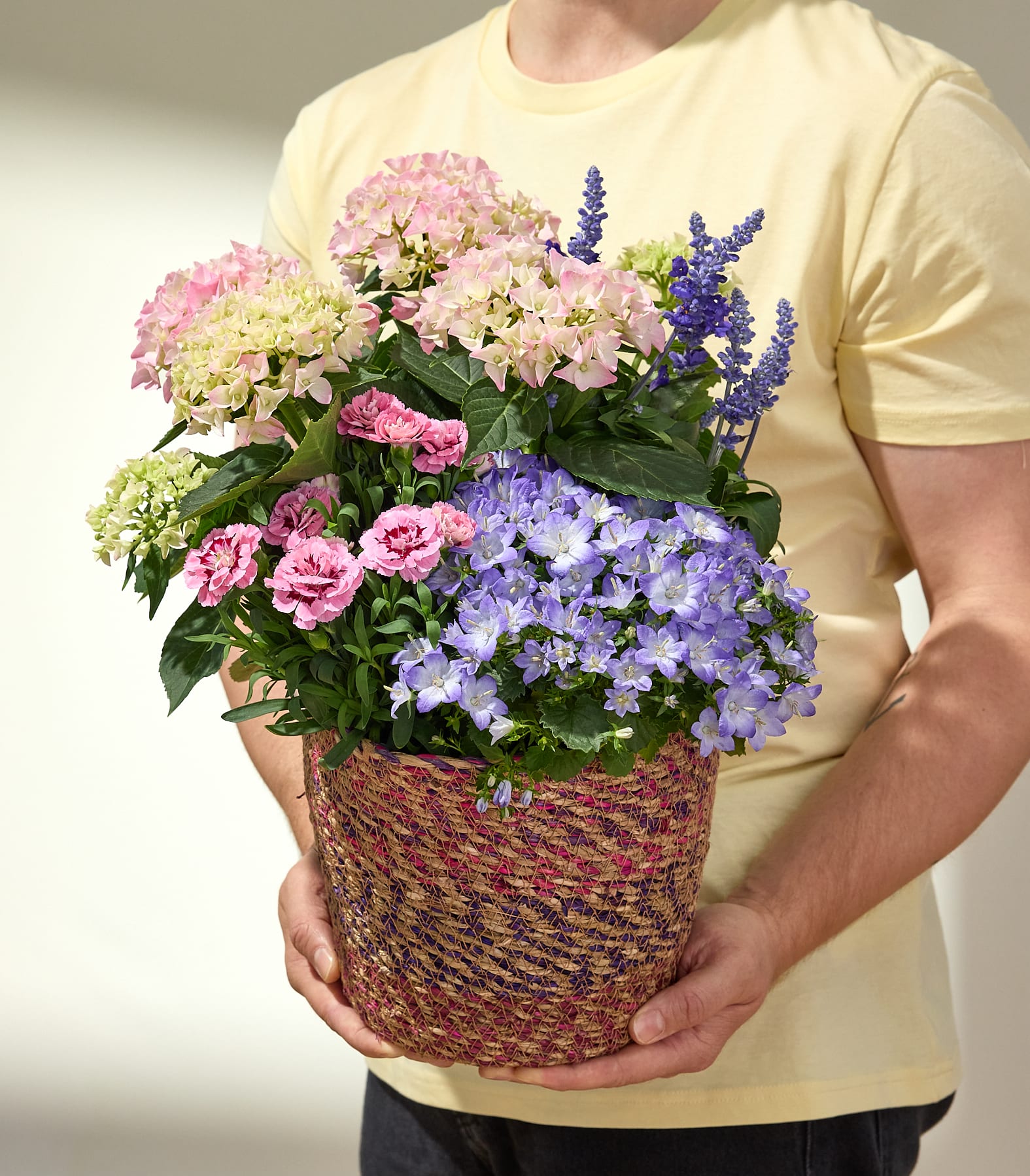 Eine Person in einem gelben Hemd hält vor einem schlichten Hintergrund einen geflochtenen Topf, der mit rosa und violetten Hortensien und blauem Salbei gefüllt ist.