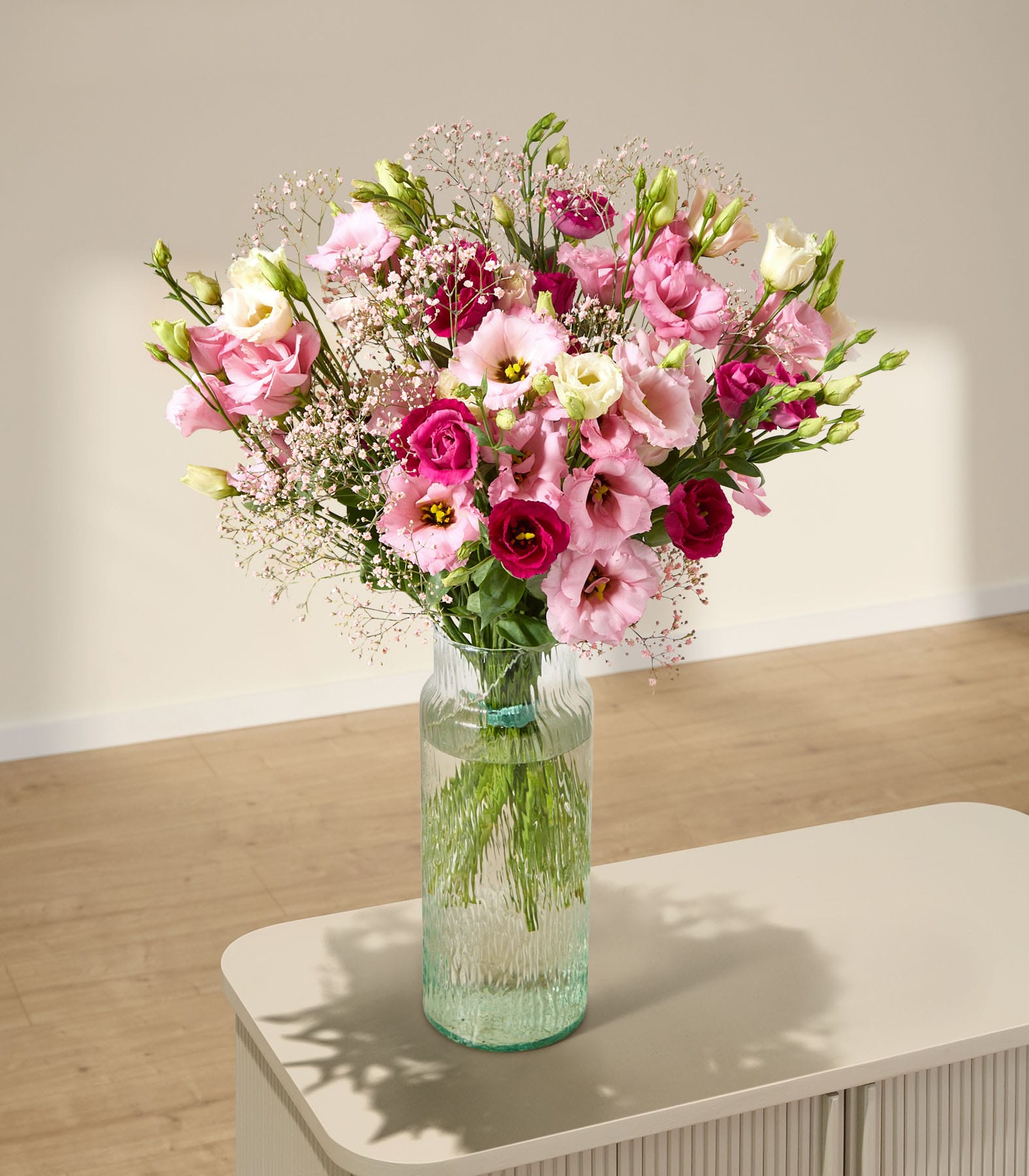 Ein farbenfroher Strauß rosa und weißer Blumen, darunter Rosen und Schleierkraut, wird vor einem schlichten grauen Hintergrund in der Hand einer Person gehalten.