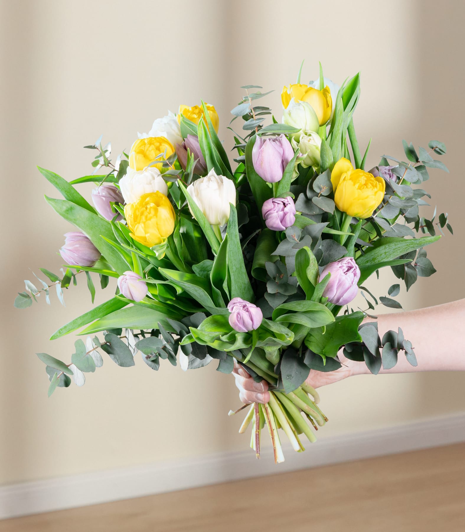 Ein leuchtender Strauß gelber und rosa Tulpen, umgeben von üppigem Grün, wird vor einem schlichten blauen Hintergrund in den Händen einer Person gehalten.