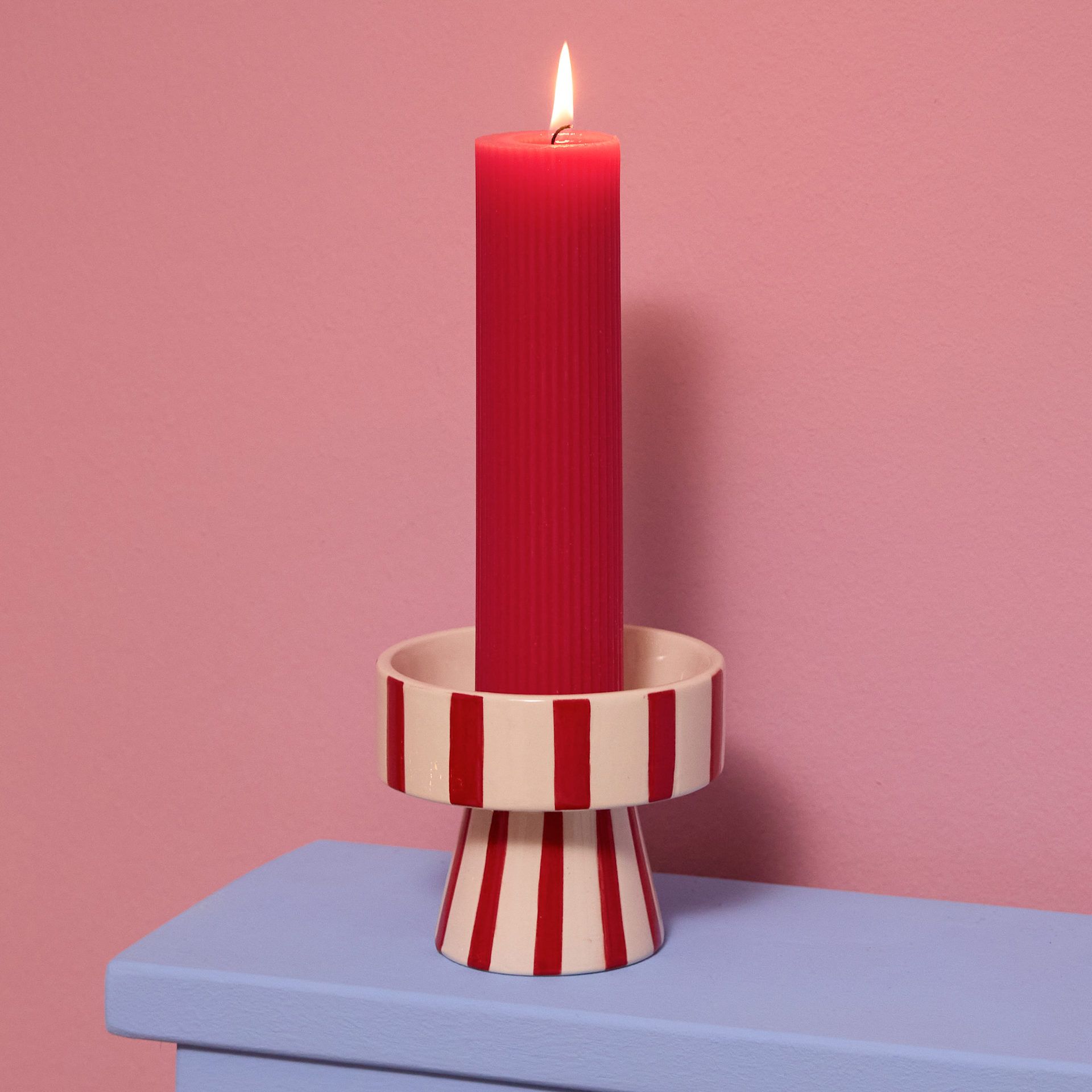 Eine leuchtend rote Kerze steht auf einem gestreiften Sockel vor einem zartrosa Hintergrund.