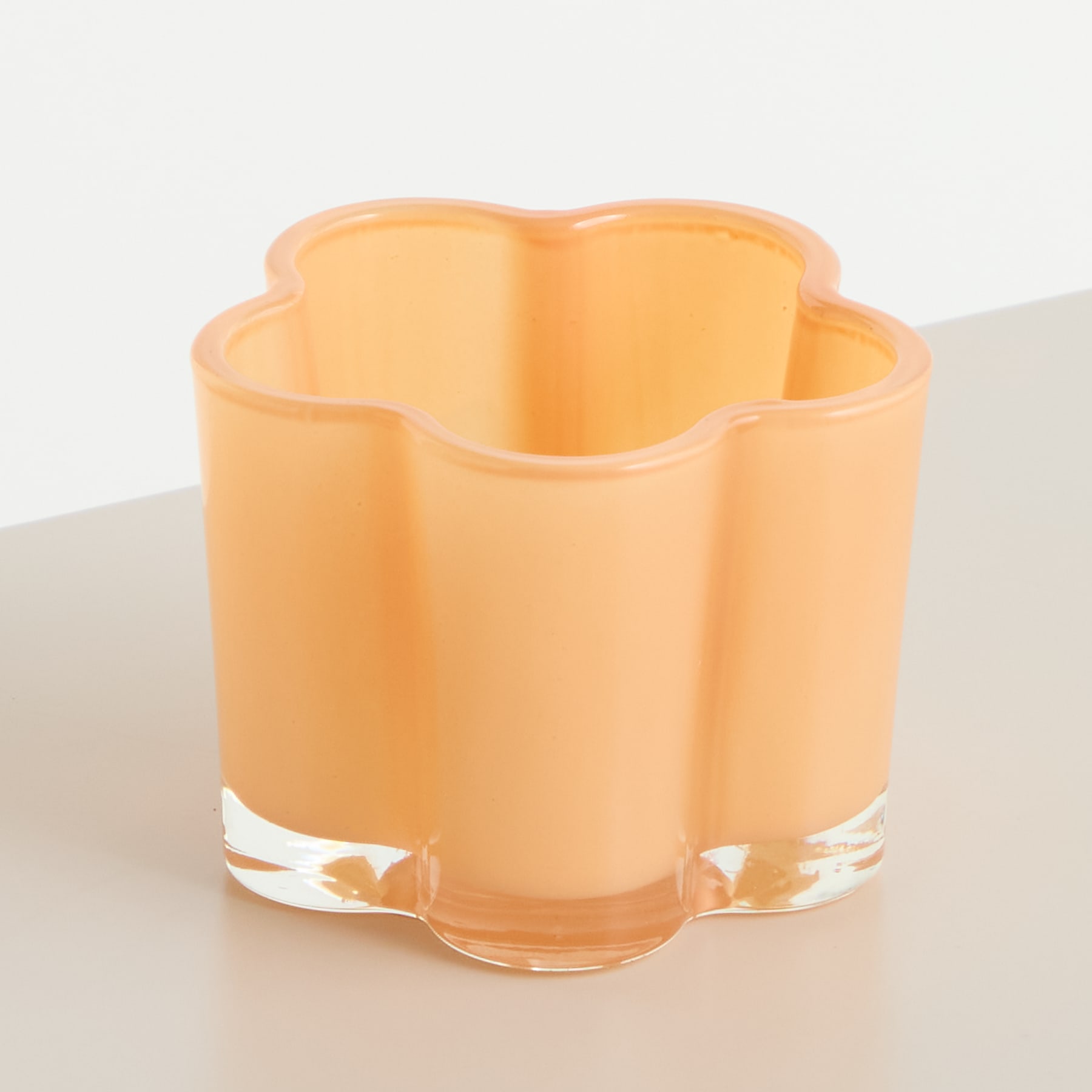 Eine zarte, pfirsichfarbene Glasvase mit Blumenform steht auf einer schlichten, hellen Oberfläche.