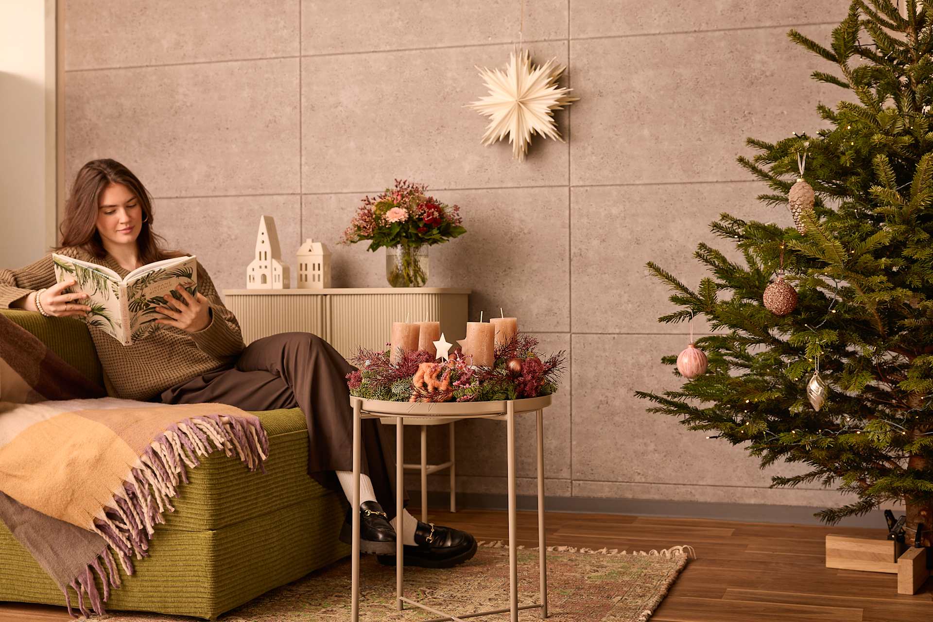 Eine Frau sitzt auf einem Sofa und liest ein Buch. Umgeben ist sie von einem geschmückten Weihnachtsbaum, Blumenarrangements und anderer festlicher Dekoration in einem gemütlichen Wohnzimmer.