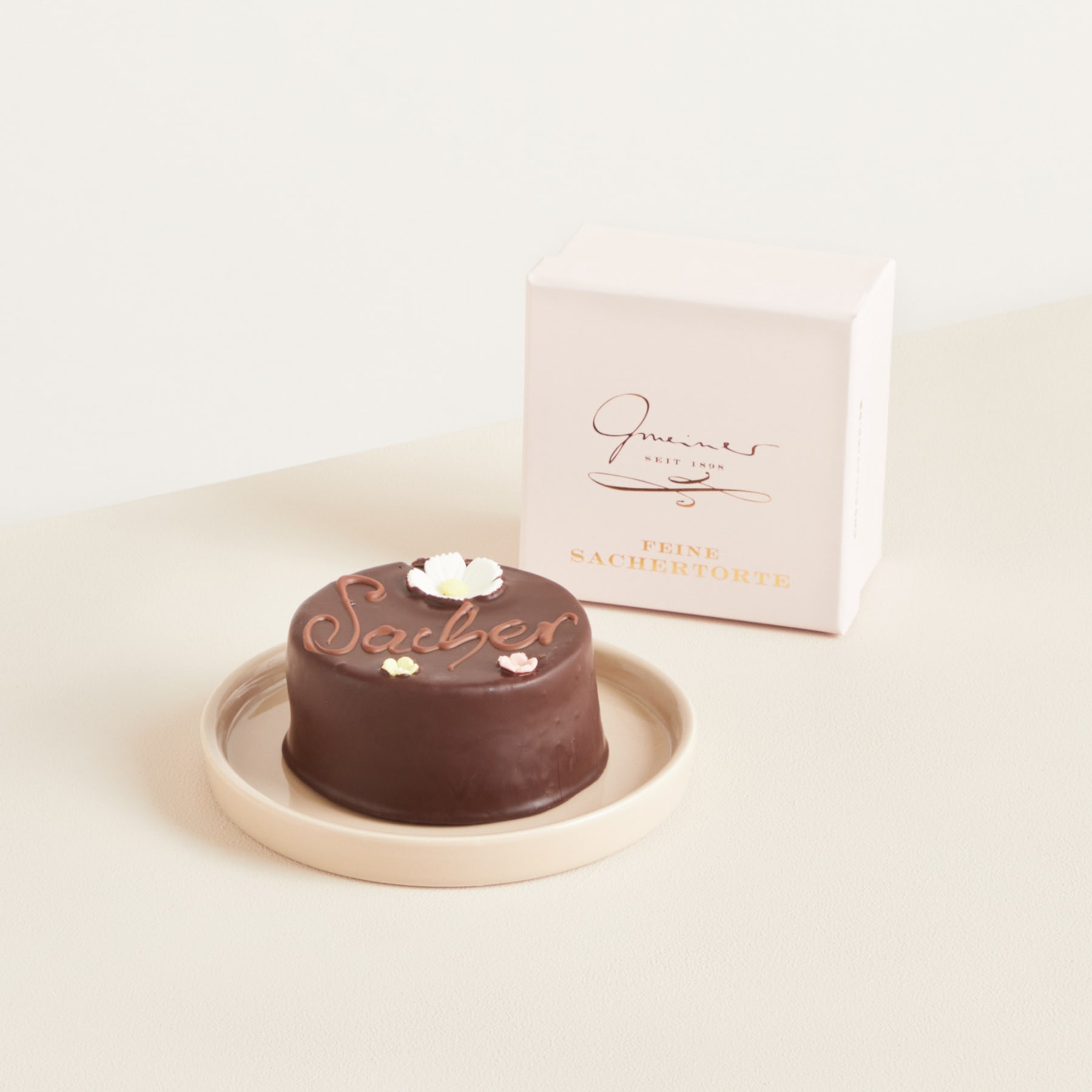 Auf einem hellen Teller steht ein kleines, rundes Schokoladendessert. Dazu gibt es eine Schachtel mit dem Aufdruck „Gourmet Chocolate“ auf einem schlichten, hellen Hintergrund.