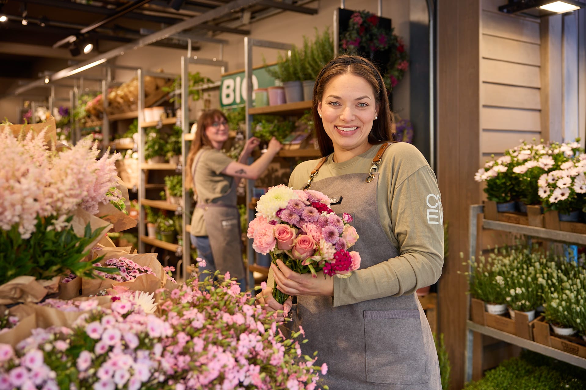 Eine lächelnde Frau in einer geblümten Schürze steht inmitten leuchtender Blumen und Pflanzen in einem Gebäude, das wie ein Blumenladen oder eine Gärtnerei aussieht.