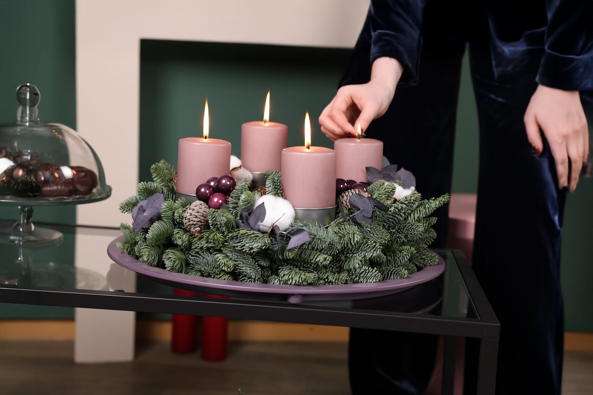 Auf einem Glastisch steht ein festlicher Adventskranz mit vier rosa Kerzen, umgeben von einem üppigen immergrünen Gesteck und dekorativen Elementen, vor einer dunkelgrünen Wand.