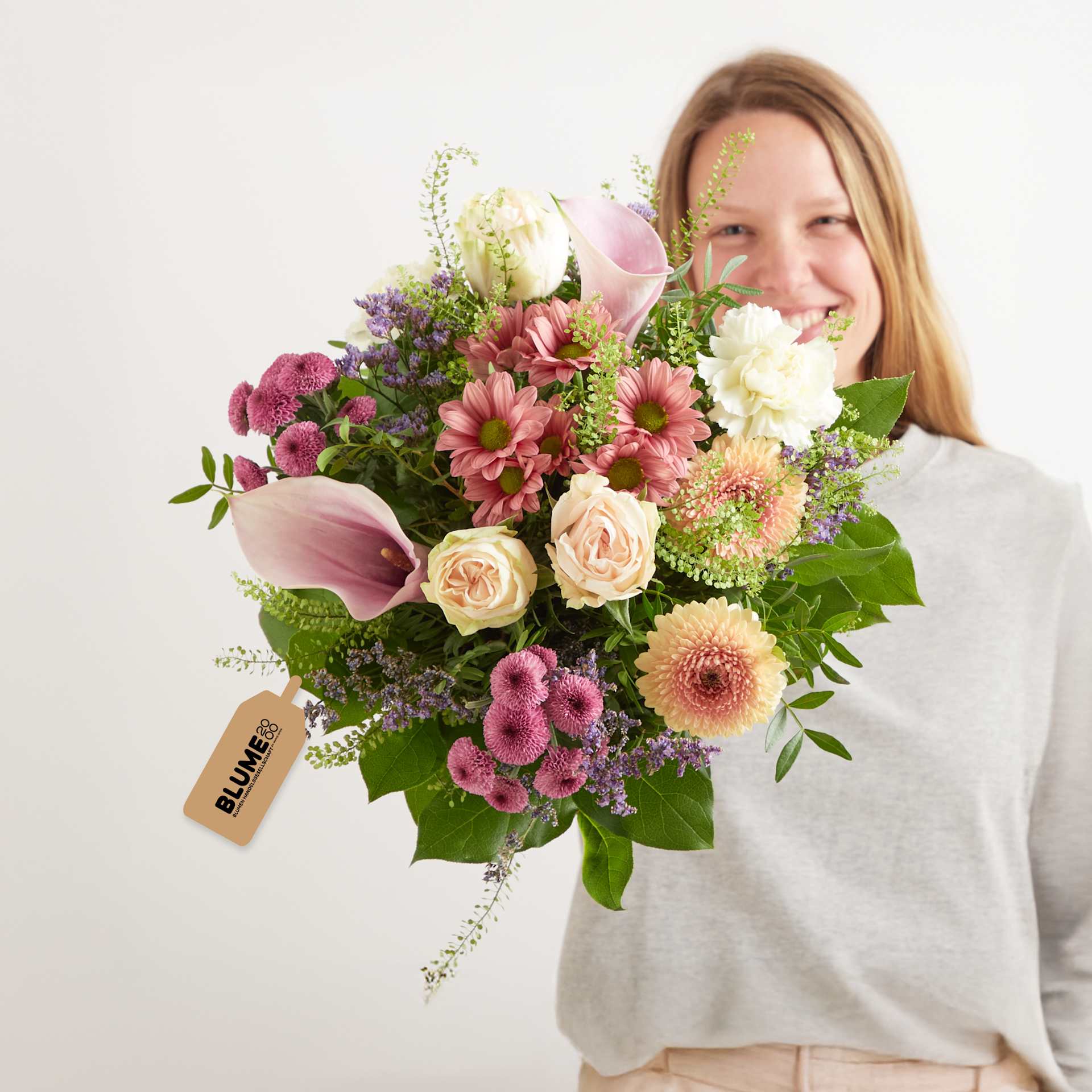 Eine lächelnde Frau hält einen farbenprächtigen Strauß verschiedener Blumen, darunter Rosen, Gänseblümchen und Dahlien, vor einem schlichten weißen Hintergrund.