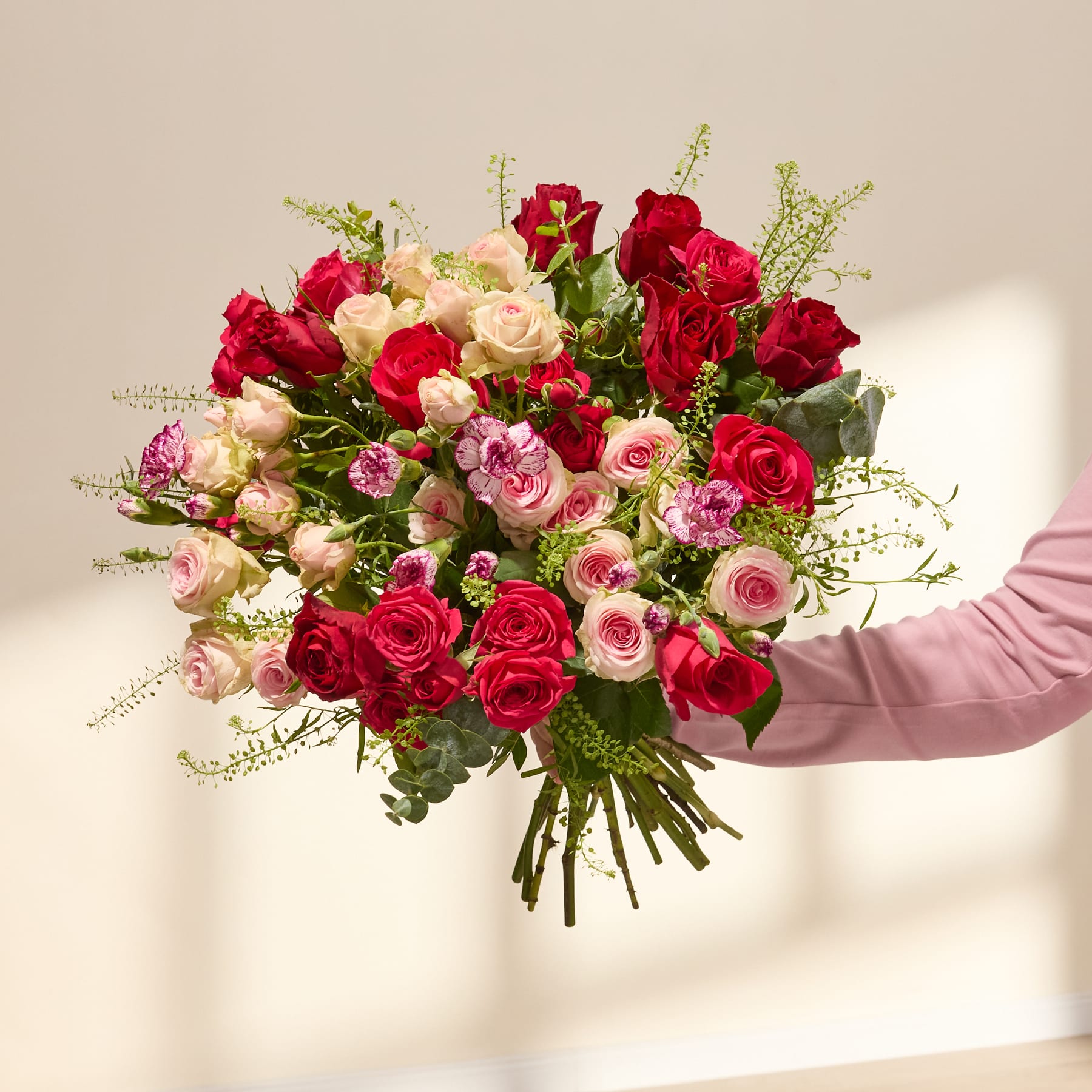 Ein großer, leuchtender Strauß roter Rosen und rosa Nelken, gehalten von einer Person in einem burgunderfarbenen Hemd vor einem schlichten weißen Hintergrund.
