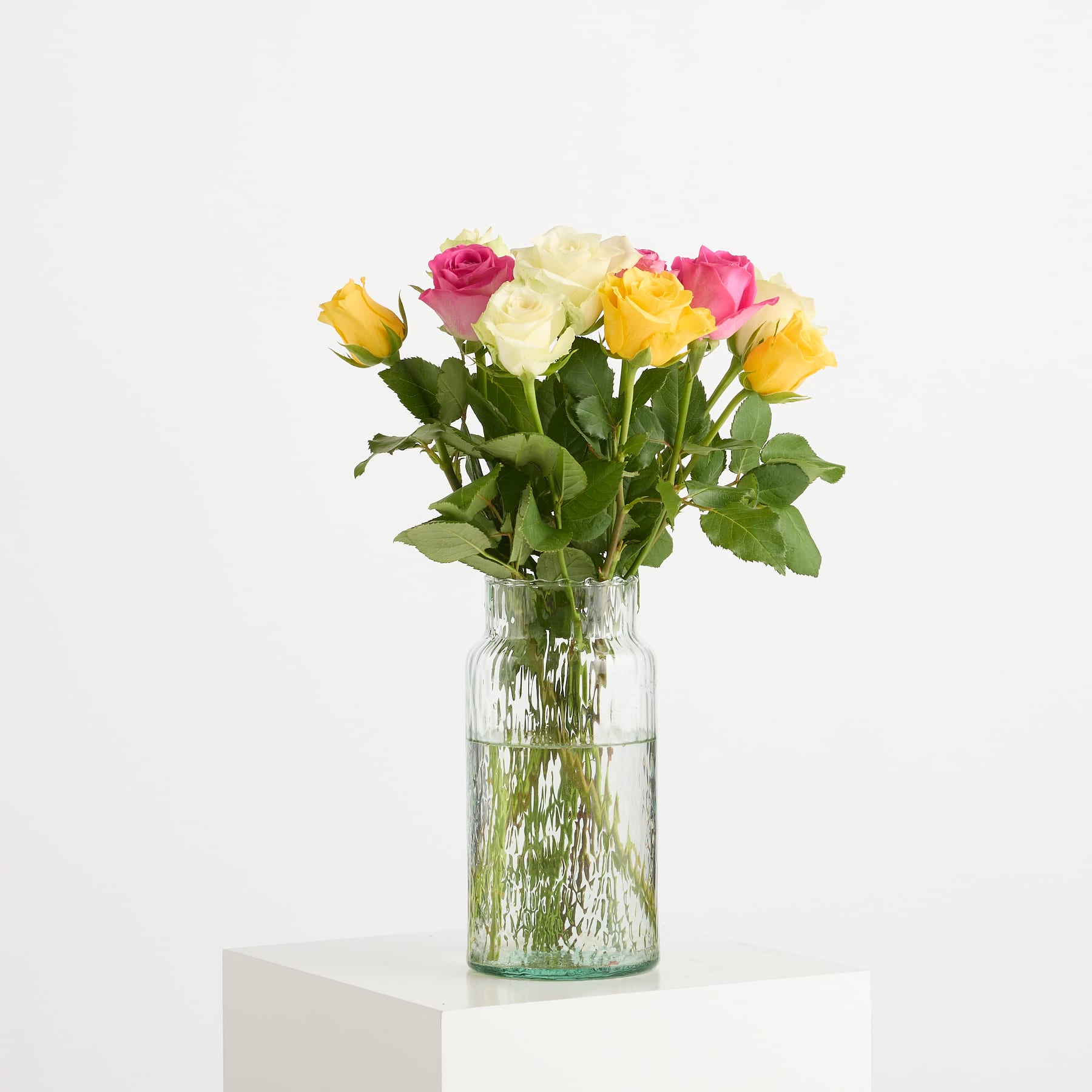 Ein Strauß bunter Rosen in einer Glasvase steht auf einer weißen Fläche vor einem schlichten Hintergrund. (Beschreibungstext KI-generiert)