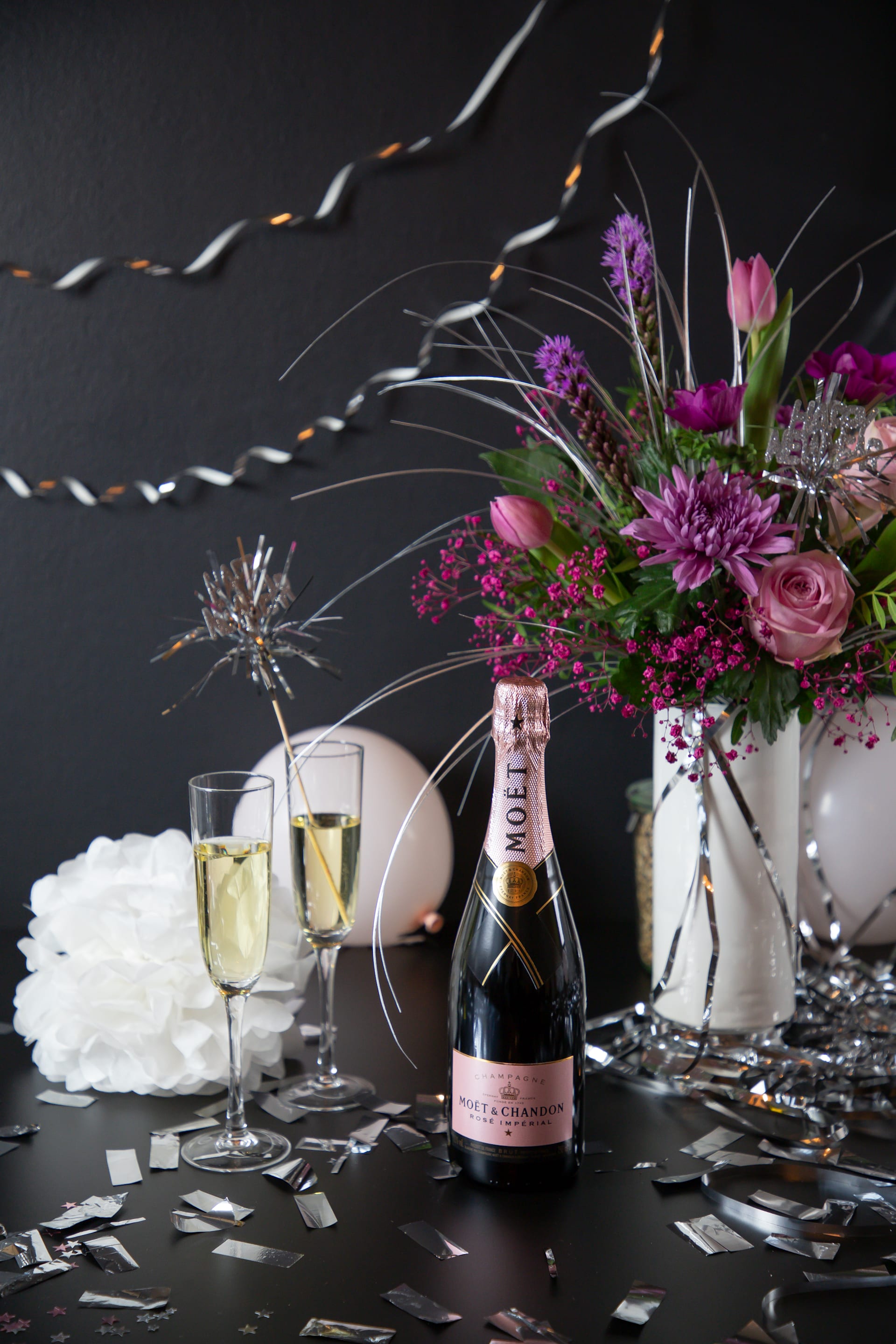 Ein festliches Arrangement mit einer Flasche Champagner, zwei Champagnergläsern und einem farbenfrohen Blumenstrauß vor dunklem Hintergrund mit verstreutem Konfetti.