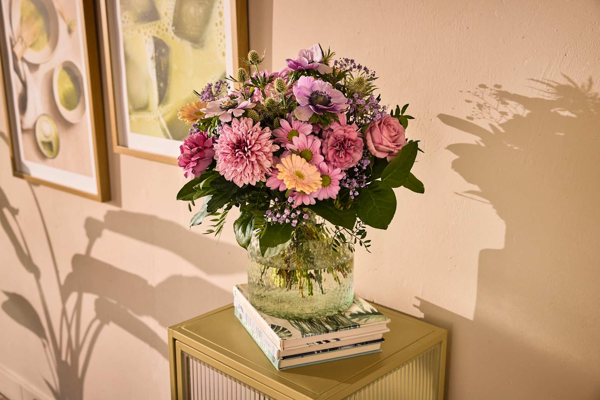 Ein leuchtender Strauß rosa und gelber Blumen thront auf einem Bücherstapel und wirft Schatten an die dahinterliegende Wand vor einem warmen, neutralen Hintergrund.