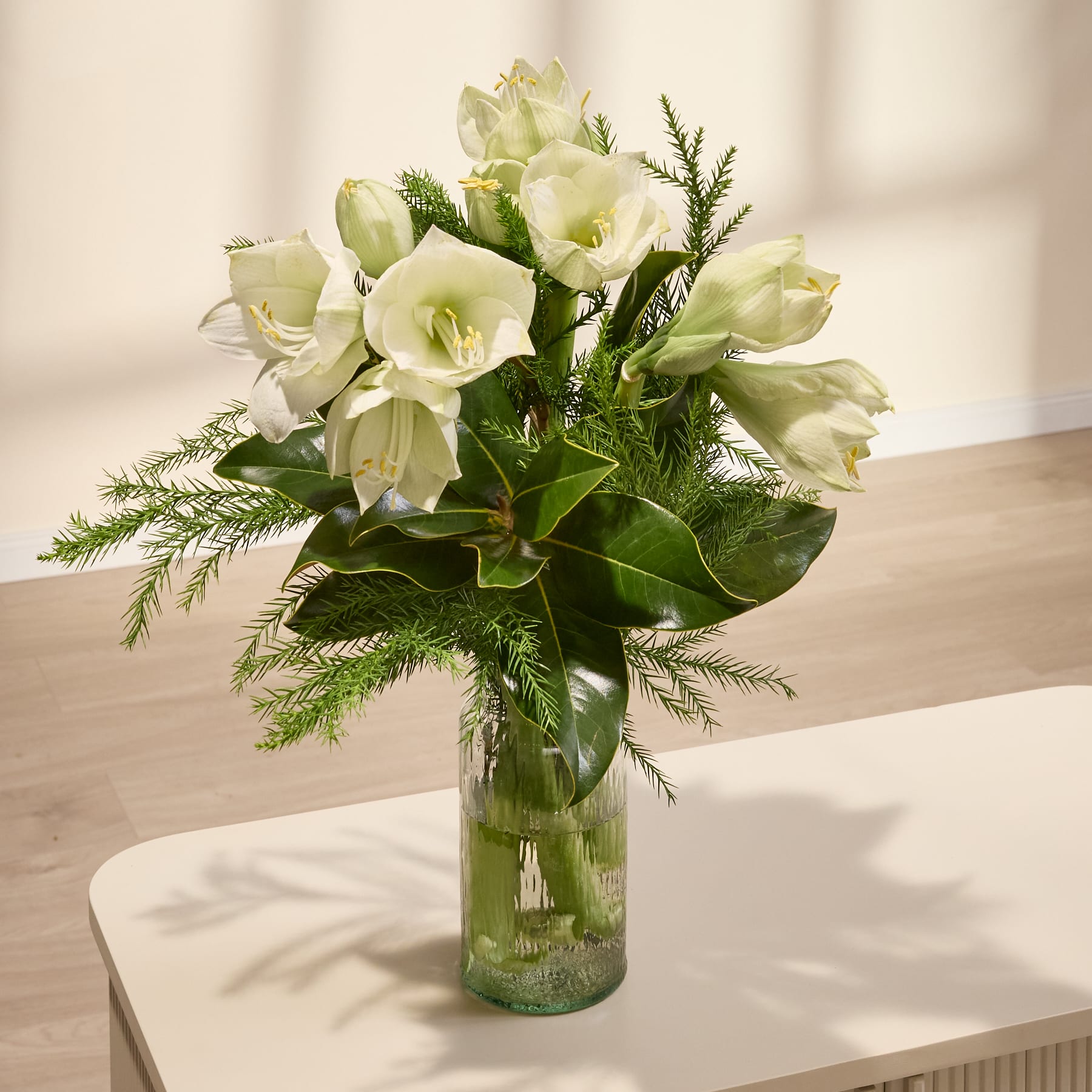 Eine Person in einem lässigen grauen Pullover hält vor einem schlichten weißen Hintergrund einen Strauß wunderschöner weißer Blumen, darunter Lilien und Grünpflanzen.